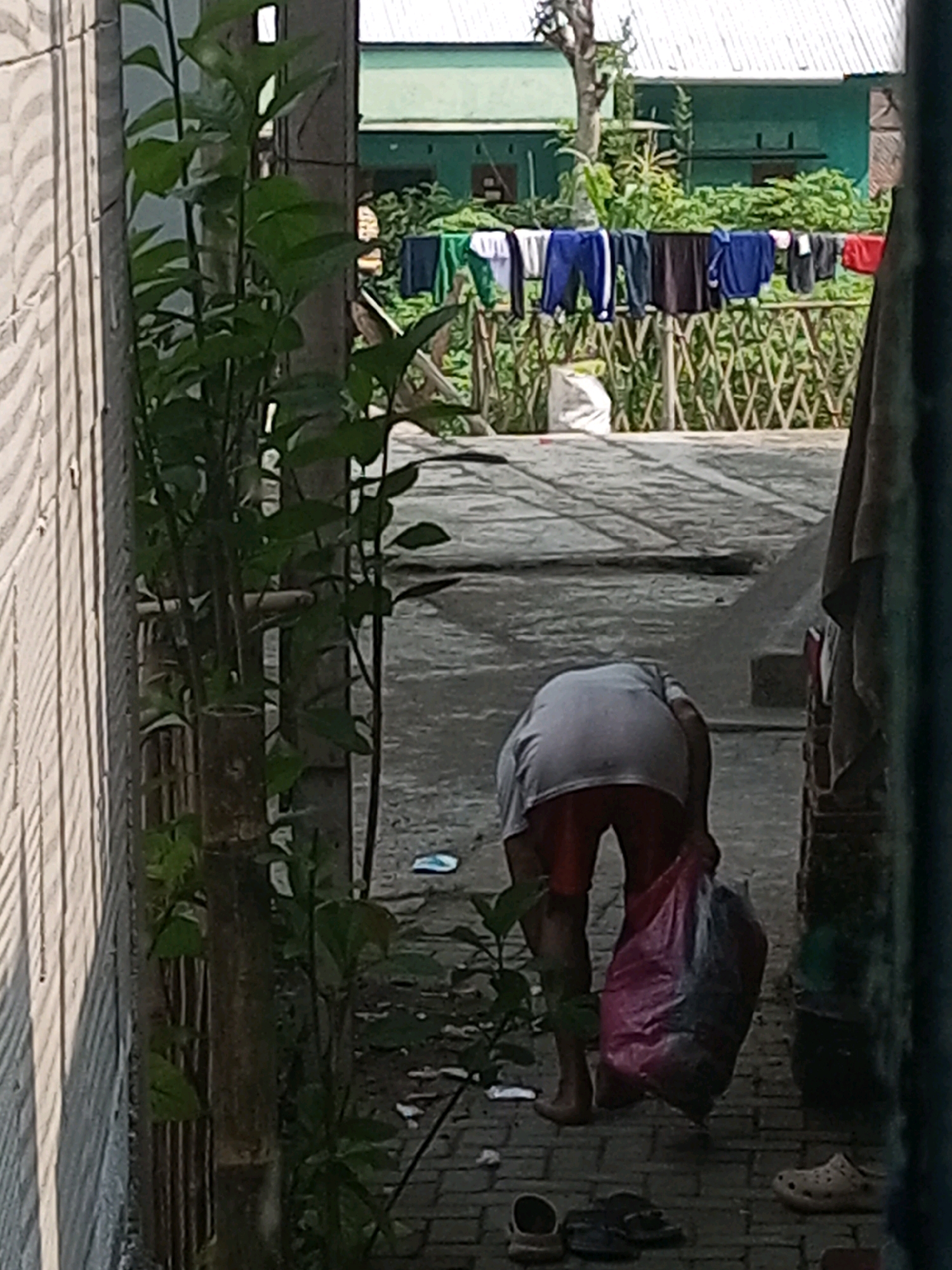 In Andongsili, Indonesia, a person is bent over, holding a large pink and blue plastic bag filled with items. The person is wearing a grey t-shirt and red shorts. They are standing on a paved outdoor surface, with a brick wall to their right and a stone wall on their left. In the background, laundry is hanging on a clothesline across a bamboo fence. Green foliage is visible throughout the scene. Several pairs of shoes, including a beige clog, are scattered on the ground.