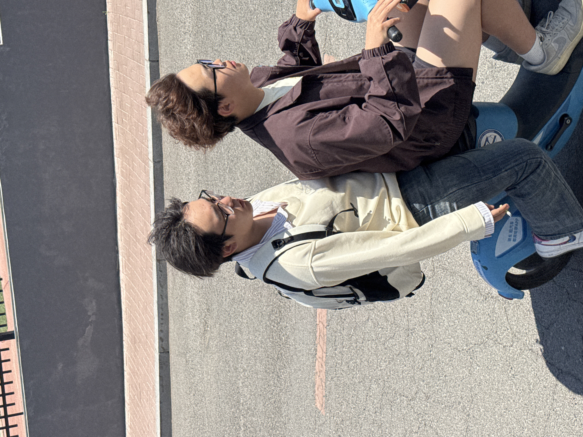 This image, rotated ninety degrees clockwise, captures two young individuals sitting on a blue shared electric scooter outdoors in Zhongtun, China, under bright sunlight.
The person in the foreground (left in the original orientation) has straight black hair and wears glasses. They are dressed in a light cream or beige jacket over a striped shirt and have a dark backpack. This individual is holding a cigarette in their right hand, which is resting on their knee.
The person behind them (right in the original orientation) has curly brown hair and also wears glasses. They are wearing a dark brown jacket over a light-colored top and are holding the handlebars of the blue scooter. Their legs are visible, with one foot wearing a light grey sneaker resting on the scooter's footrest.
The scooter itself is bright blue and features Chinese text. Visible text includes "共享电动车" (gòngxiǎng diàndòngchē), meaning "shared electric vehicle/scooter," and "租用电驴APP" (zūyòng diànlǘ APP), which translates to "rent electric donkey APP."
They are positioned on a paved surface, likely a path or road. To the left (top in the original orientation) is a darker gray surface bordered by a lighter, brick-patterned strip. Further left, a glimpse of green foliage is visible. The bright sun casts clear shadows, suggesting it is daytime with clear weather. The individuals appear relaxed, taking a break on the shared scooter.