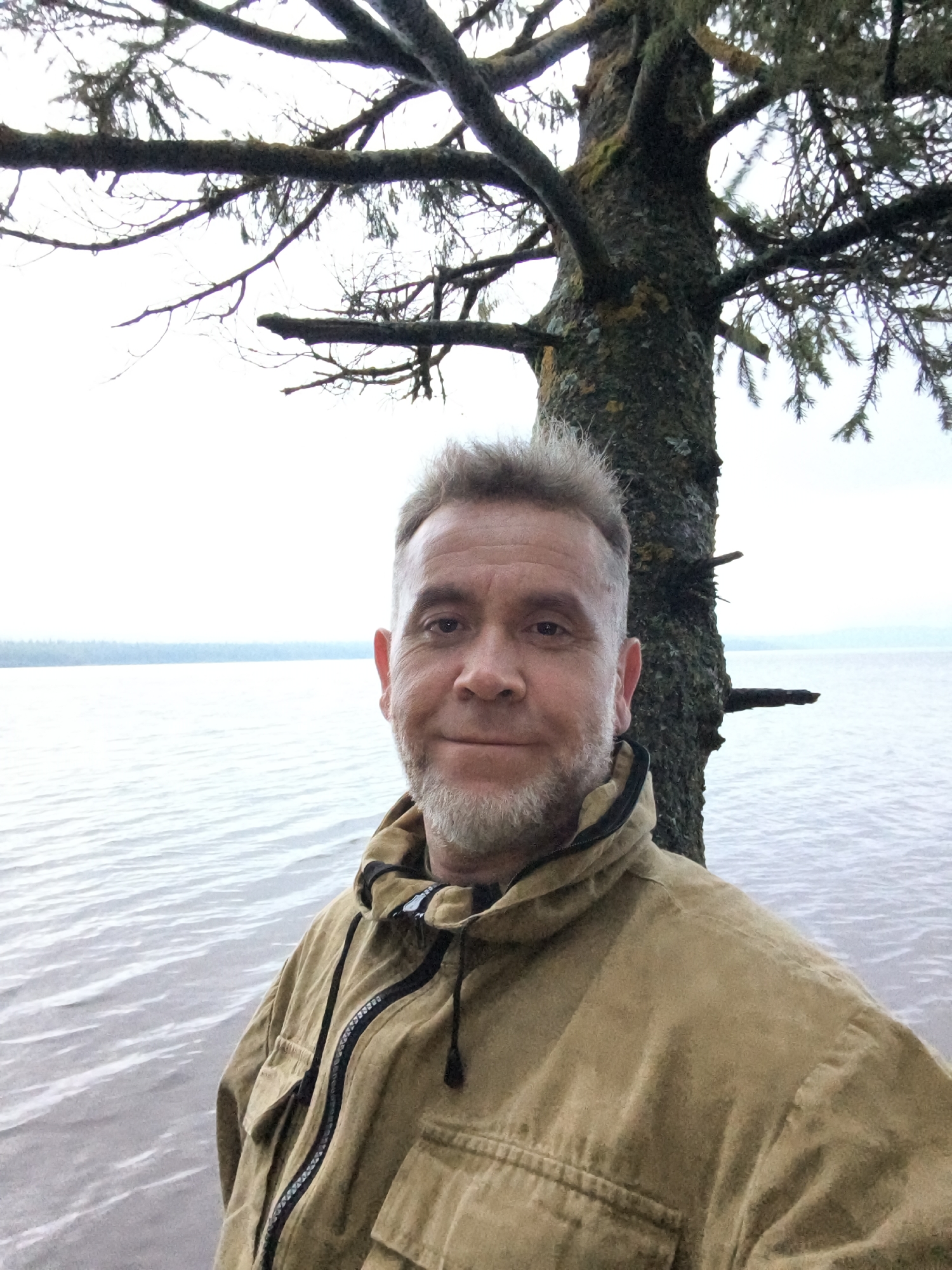 A man with short gray hair and a beard, wearing a tan jacket, stands in front of a tree. The tree is tall and rough-barked with moss. Behind the tree is a body of water with ripples and a faint horizon line. The sky is overcast. The man is looking at the camera and smiling. The location is Zyuratkul' National Park, Russia.