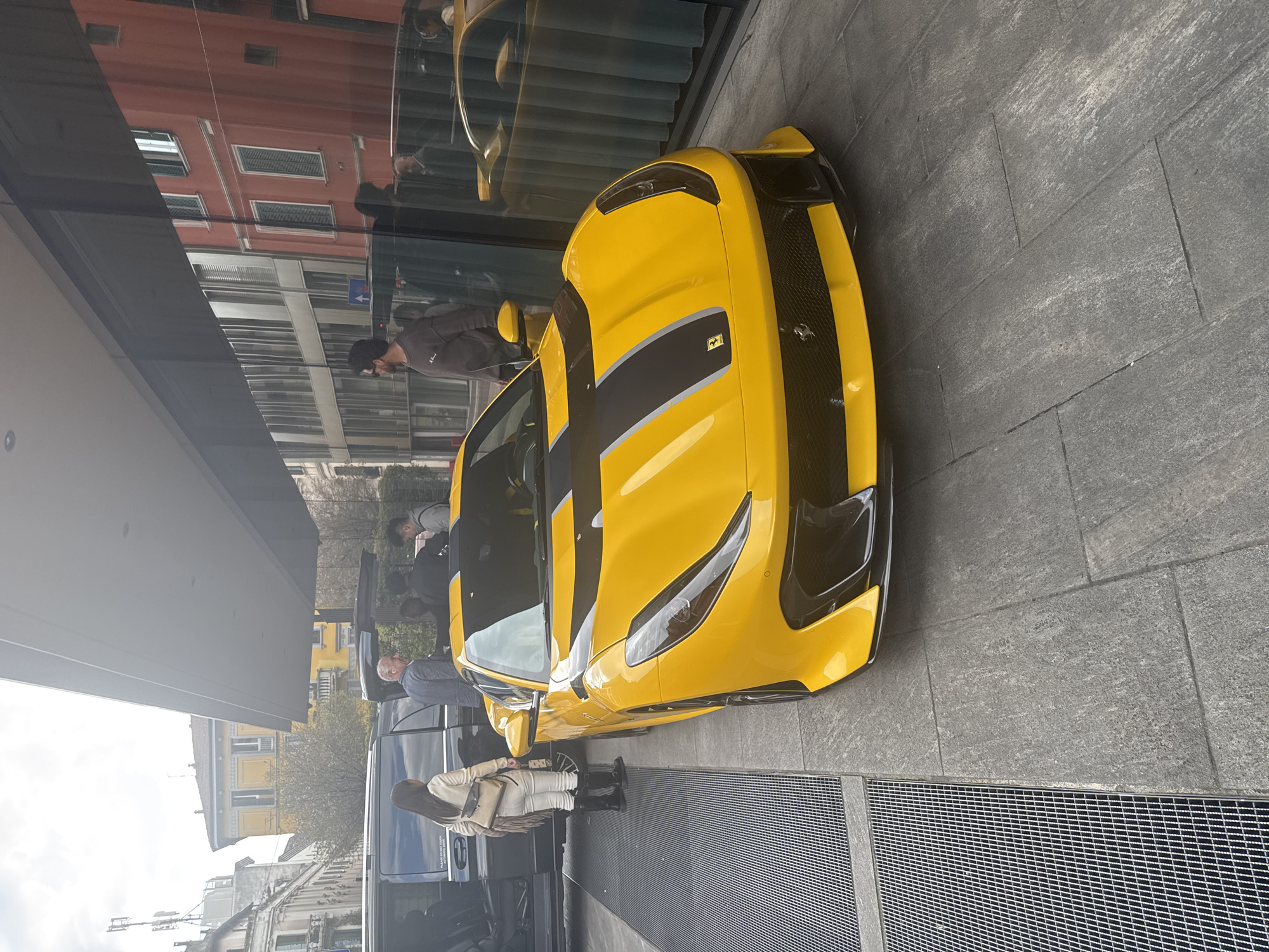 A yellow Ferrari sports car, identified as a Ferrari 812 Competizione with twin black racing stripes on its hood, is parked on grey rectangular paving stones in an urban setting. The car is positioned in front of a modern building with a dark glass facade, which reflects a reddish-orange classical building and a contemporary structure across the street. An overhead white, flat canopy extends from the modern building, sheltering the area.
To the left of the Ferrari, a dark grey or black luxury minivan (likely a Mercedes-Benz V-Class or similar) has its passenger side sliding door open. A woman with dark hair, wearing a light-colored jacket, white trousers, and dark boots, carrying a beige shoulder bag, stands near the open door of the minivan, facing away from the Ferrari. A grey-haired man in a light grey shirt stands between the Ferrari and the minivan, looking towards the left with a slight smile. Two other men are partially visible near the minivan's open door.
To the right of the Ferrari, a man with dark hair and a beard, wearing a dark grey long-sleeved shirt and sunglasses, stands looking towards the left. His reflection is visible in the glass facade behind the car. The ground consists of large grey paving stones, with a section of metallic grates visible to the far left of the Ferrari. In the background, beyond the vehicles, older, multi-story European buildings, including one yellow and one light-colored, are visible under an overcast sky. A white antenna structure is present on a distant rooftop. The scene takes place in a bustling, upscale area of Milan, Italy.