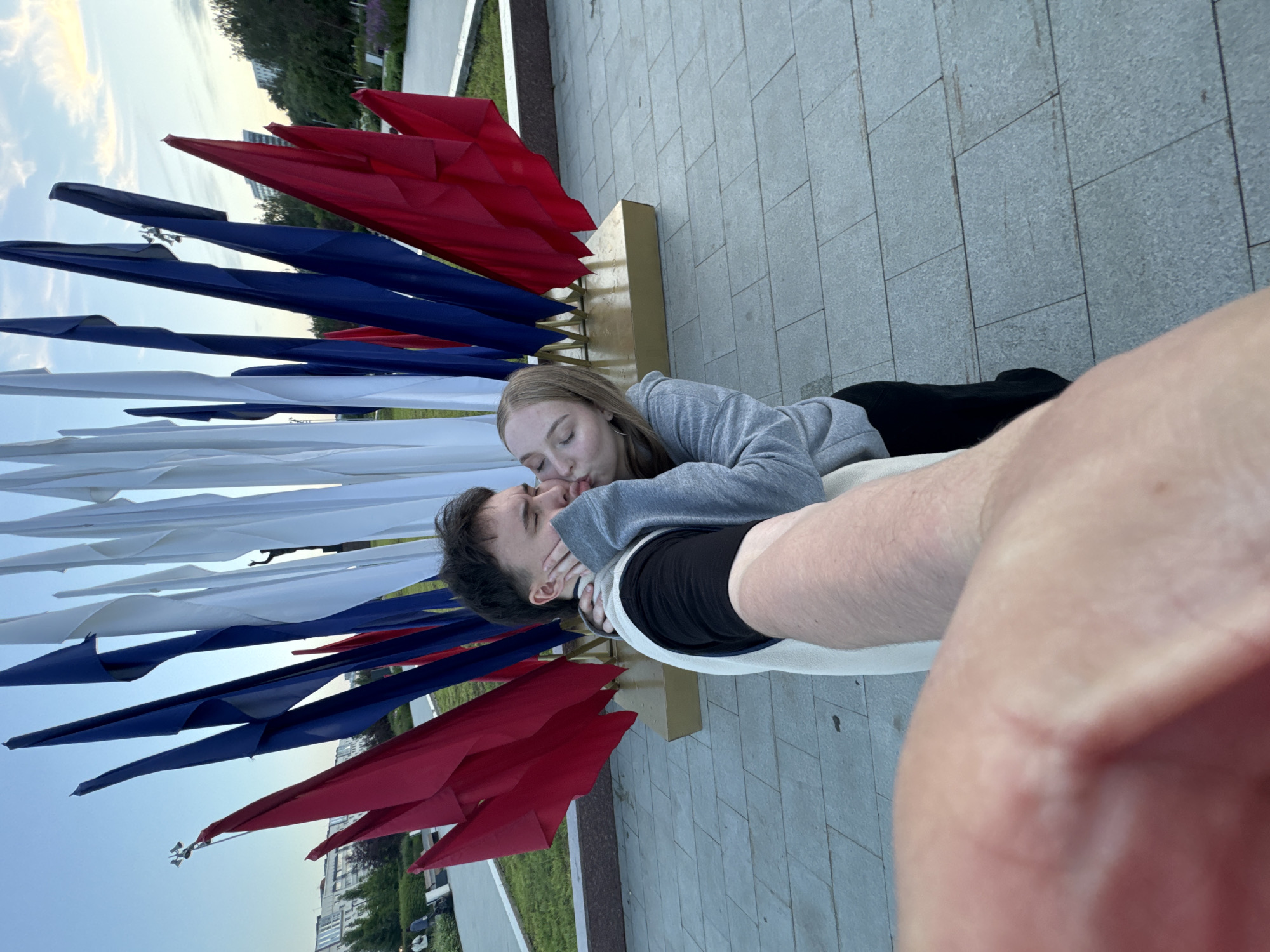 A couple is captured in a selfie, embracing and kissing. The woman, with blonde hair, wears a grey hooded sweatshirt and a black skirt. The man, with dark hair, is dressed in a black t-shirt and a white vest. They are positioned in front of a display of red, white, and blue flags, which are arranged in a fan-like pattern. Behind the flags, a silhouette of a person is visible, as well as some buildings in the distance. The ground is paved with large, grey square tiles. The sky is clear with a hint of sunset colors. The image appears to have been taken in a public outdoor space, possibly a park or monument area. Based on the context of the flags, the location is likely in the city of Perm, Russia.