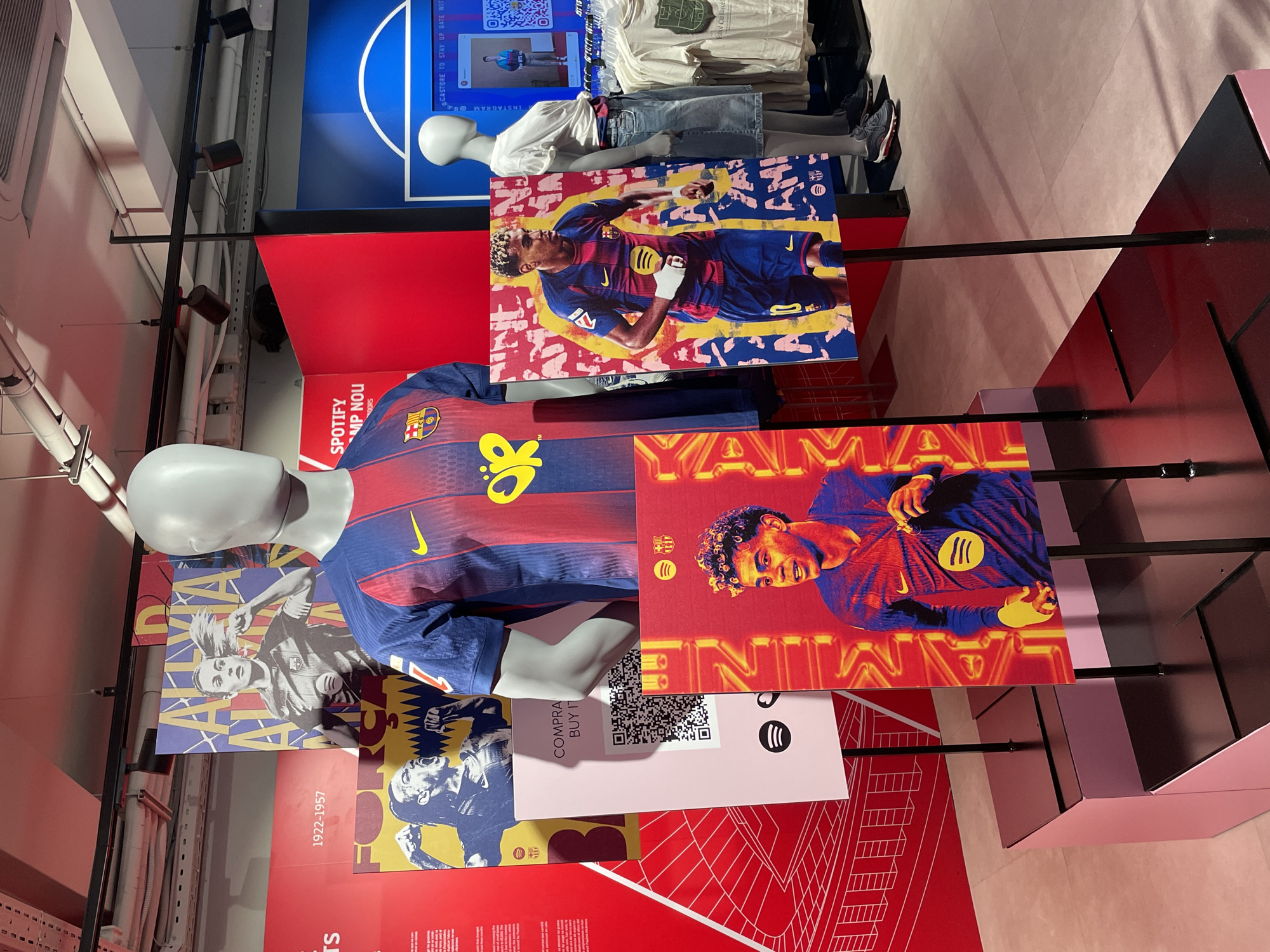 The image shows an indoor display, possibly a store or exhibition in Barcelona, Spain, featuring FC Barcelona merchandise and promotional material. Several mannequins are present, one wearing a Barcelona jersey with a large "10" on the front. There are also large posters or prints of Barcelona players, including one that appears to be of "YAMAL". The environment suggests a commercial or promotional space designed to showcase and sell Barcelona-related items. The color scheme is dominated by the club's colors of blue, red, and yellow. A QR code is visible, likely for purchase information. Other clothing and promotional materials are visible in the background. The lighting is bright and well-distributed.