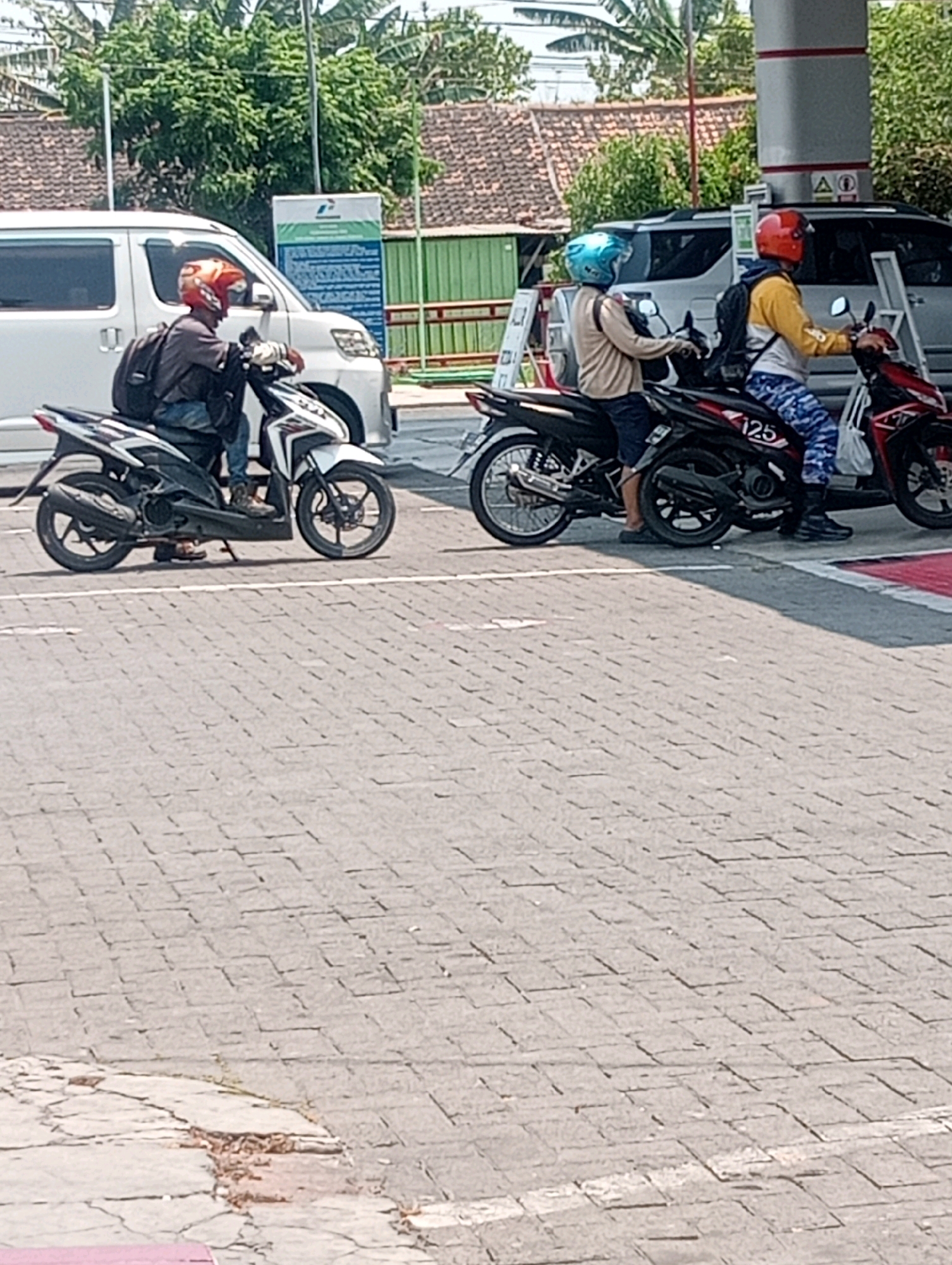 In Plosojenar, Indonesia, three individuals are observed at a gas station, each on a motorcycle. The first individual, situated on the left, is wearing a dark shirt and jeans, with a red helmet and a black backpack. Their white and black motorcycle is positioned on a paved surface. To the right of this individual, a second person in a light brown shirt, dark shorts, and a blue helmet rides a black motorcycle. Further to the right, a third individual sports a yellow and white shirt, blue and white patterned pants, and a red helmet, also on a black motorcycle. Behind the motorcycles, a white van is visible, alongside a green fence and a building with a tiled roof. The ground is paved with interlocking grey bricks.