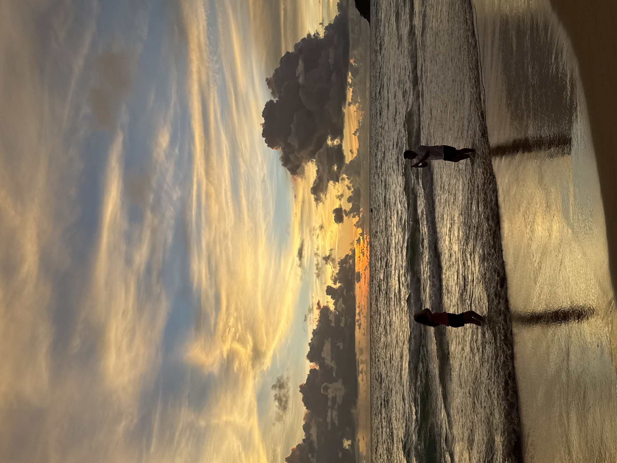 The image depicts a serene sunset scene on a beach in Karon, Thailand. The sky is a canvas of warm hues, with streaks of orange, yellow, and blue clouds stretching across the frame. Dark, dramatic clouds are scattered amongst the lighter ones, creating a sense of depth and contrast. The ocean reflects the vibrant colors of the sky, with gentle waves lapping at the shore.
Two individuals are visible in the water, silhouetted against the bright sunset. One person is in the foreground, appearing to be wading or standing in the shallow water. Further back, another person is also in the water. Their presence adds a human element to the breathtaking natural scenery.
The time of day is clearly sunset, as indicated by the rich colors in the sky and the long shadows cast on the water. The weather appears to be calm, with the gentle waves suggesting a peaceful evening. The location cues point to a beach setting, with the vast expanse of water and the sandy shore. The overall emotion conveyed by the image is one of tranquility and awe, capturing the beauty of nature at its finest. No visible text is present in the image.