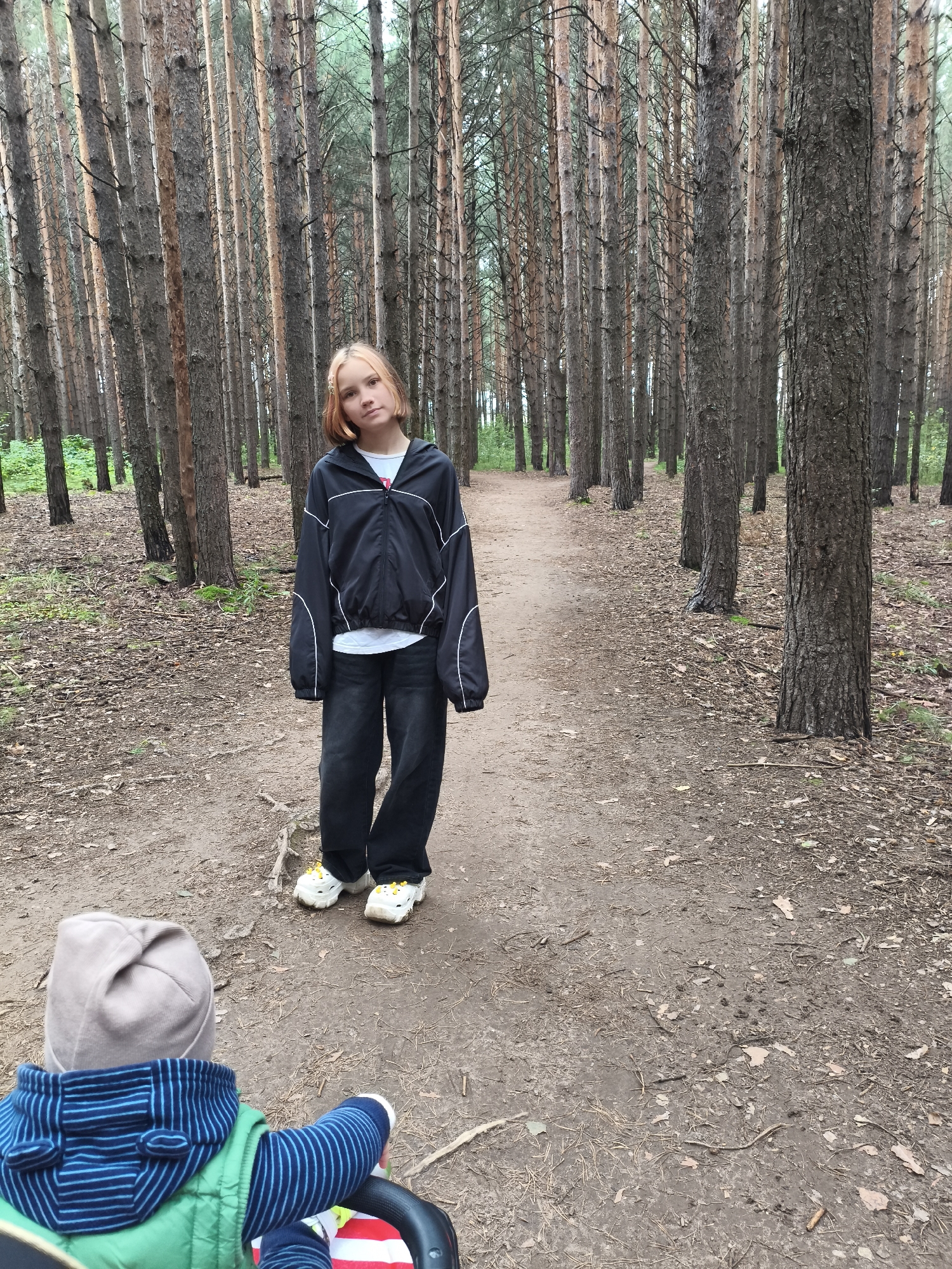 In a pine forest in Chaikovsky, Russia, a young girl stands on a dirt path, facing the camera. She has short, blonde hair with reddish highlights and is wearing a black zippered jacket with white piping over a white t-shirt, and dark, baggy jeans. She is also wearing white sneakers with yellow accents. In the foreground, a child in a green vest and a blue and striped hooded jacket is facing away from the camera, seated in a stroller. The path is lined with tall pine trees, and the ground is covered with pine needles and fallen leaves. The sky is overcast.
