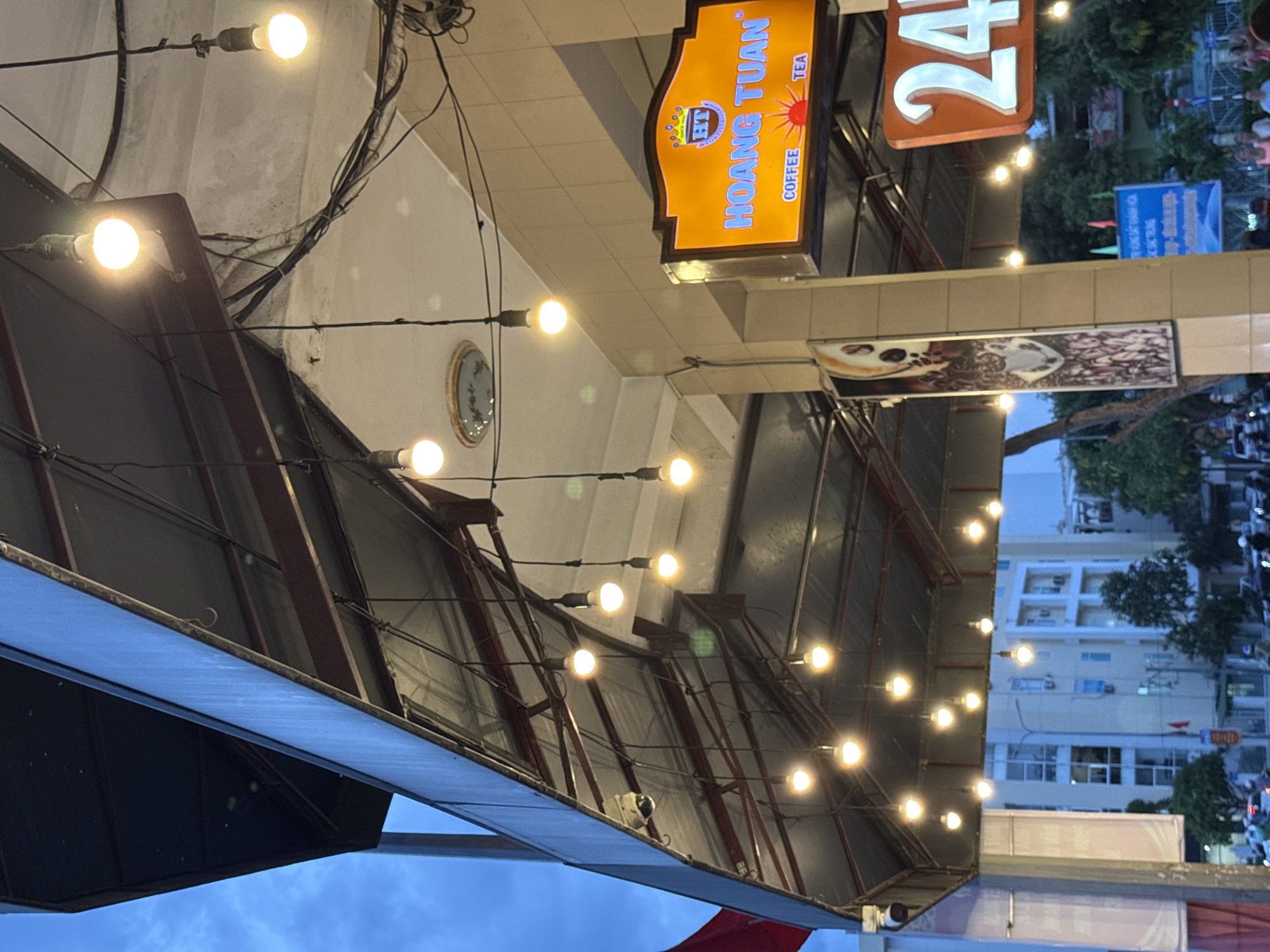 The image depicts the exterior of a coffee shop in Nha Trang, Vietnam, as indicated by the sign reading "HOANG TUAN COFFEE TEA". The shop has an overhang supported by metal beams, from which numerous pendant lights are suspended. The lights are illuminated, casting a warm glow. A security camera is visible mounted on the underside of the overhang. In the background, buildings and trees are present, suggesting an urban environment. The sky is overcast. The image is taken from a low angle, looking up at the overhang and lighting.