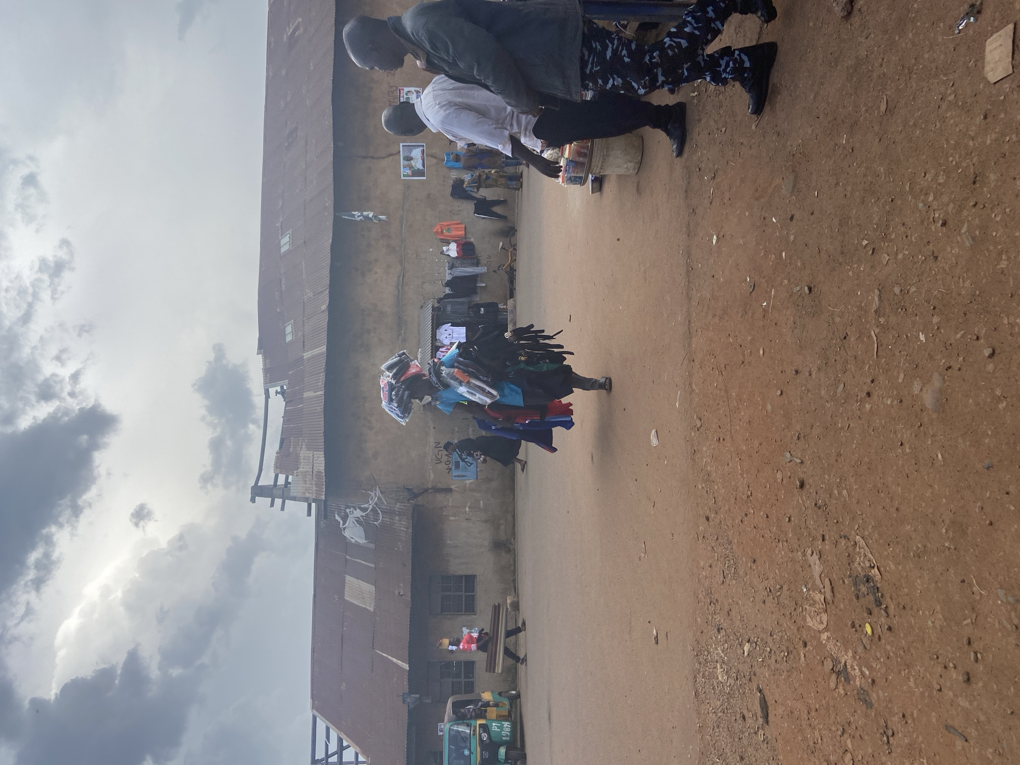The image depicts an outdoor scene in Bukuru, Nigeria, likely a market or a street stall. The foreground is dominated by a dusty, reddish-brown dirt ground, with some scattered debris. On the right side of the frame, a rusty metal roof of a building runs along the length of the image.
Several people are present. In the upper part of the image, two men are visible. One is wearing a dark jacket and the other a white shirt, both appearing to be in conversation. Further down, a person is carrying a stack of colorful clothing items, possibly for sale. Another individual is seen in the mid-ground, near a wall with several windows. In the lower part of the image, a green auto-rickshaw, a common mode of transport in Nigeria, is parked on a paved surface. A person wearing a red shirt is seated on a wooden bench.
The overall setting appears to be somewhat dilapidated, with weathered buildings and a rustic ground surface. The sky is overcast with grey clouds, suggesting a potentially cloudy or impending rainy day. The image is oriented vertically, with the buildings and ground running from top to bottom.
No discernible text is visible in the image, other than what might be markings on the auto-rickshaw which are too small to read. The scene suggests a busy, informal commercial environment.