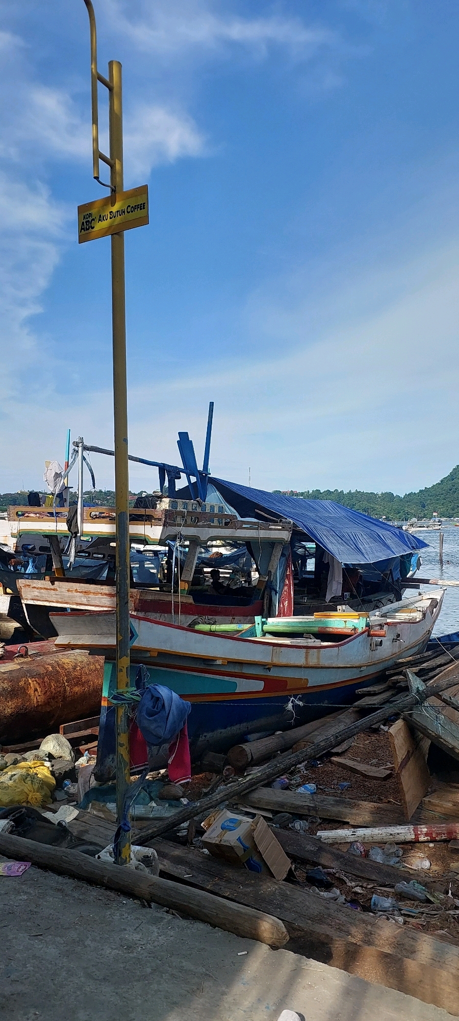 A weathered yellow sign with black lettering reading "KOPI ABC Aku Butuh Coffee" is attached to a yellow lamppost in the foreground, partially obscuring the view of boats docked in a waterfront area. The boats appear to be in a state of disrepair, with a blue tarp covering part of the structure of one boat. The surrounding area is cluttered with debris, including wooden planks, plastic bottles, and other refuse. The scene is set against a backdrop of a blue sky with scattered clouds and a distant, tree-covered hillside. The image suggests a location in Jayapura, Indonesia, given the prevalence of such signage and waterfront scenes in the city.