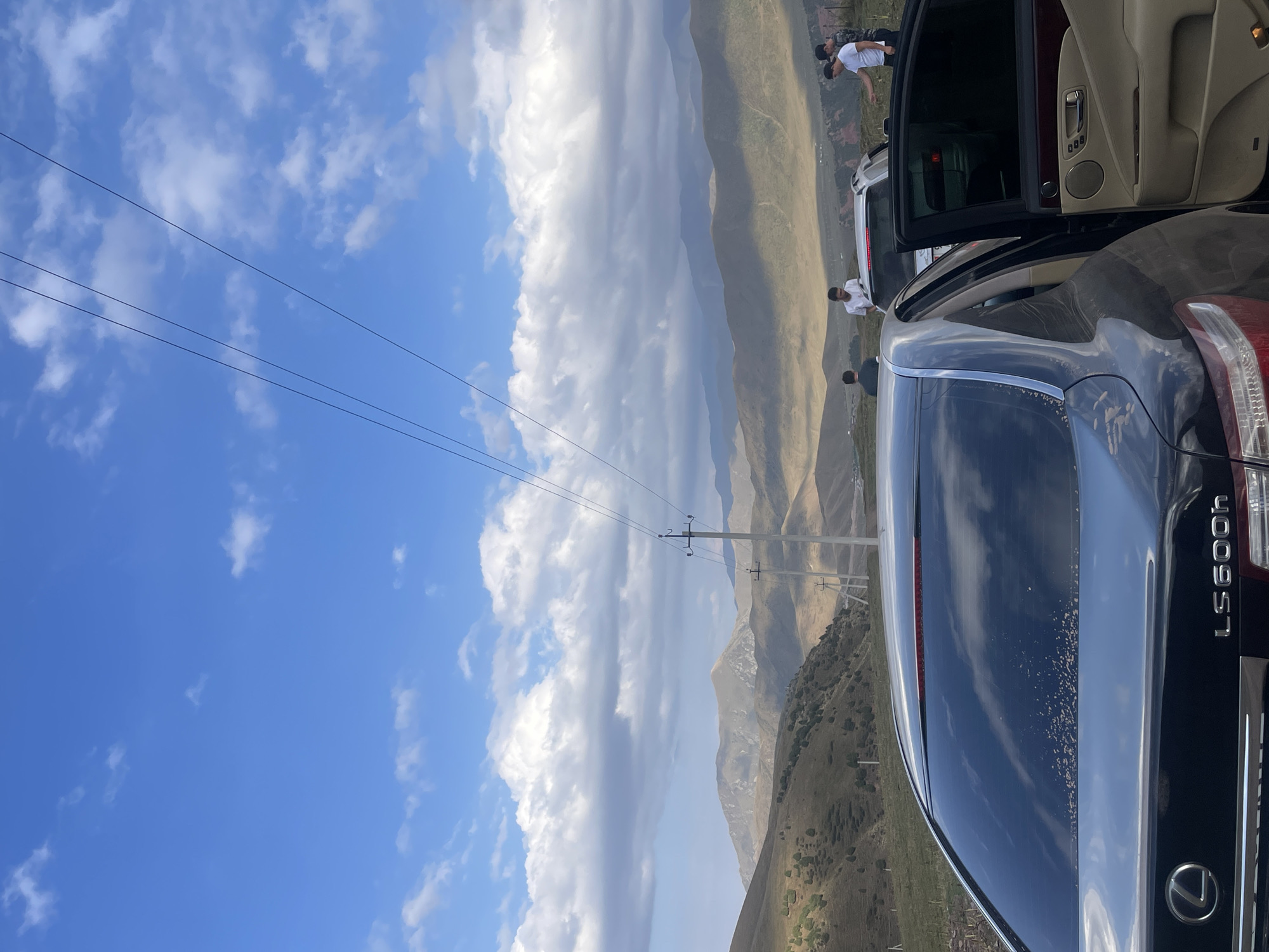 The image depicts a mountainous landscape in Chunkurchak, Kyrgyzstan, under a bright blue sky with scattered white clouds. In the foreground, the rear of a dark-colored Lexus LS600h sedan is visible, with its trunk slightly open. To the right, the driver's side door of the Lexus is also open, revealing a tan interior. Several individuals are present, interacting near the vehicles. One person is standing near the open trunk, and three others are gathered to the right of the car. In the middle ground, a white SUV is parked. The background features rolling hills and rugged mountains, with several utility poles and power lines traversing the scene. The terrain is sparsely covered with vegetation and scattered trees. The overall atmosphere suggests a stop or gathering in a scenic, rural location.
