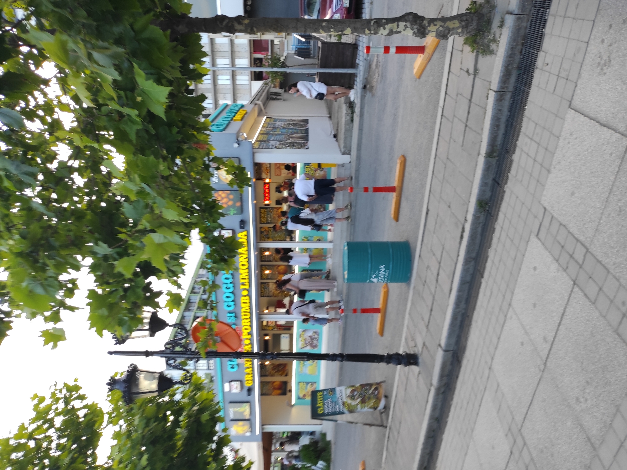 The image shows a street scene in Năvodari, Romania, with a food stand and people. The food stand appears to be selling drinks and possibly ice cream, with signage indicating "CIFFF SI GOGO" and "GRANITA PORUMB LIMONADA". Several people are gathered around the stand, some ordering and others waiting. In the foreground, a sidewalk with patterned paving stones is visible, along with a black lamppost. A blue cylindrical bin is positioned on the side of the road. Traffic cones with orange bases are placed on the road, possibly indicating a temporary restriction or safety measure. The background shows a multi-story building. The overall impression is of a casual, daytime scene in a commercial area.
