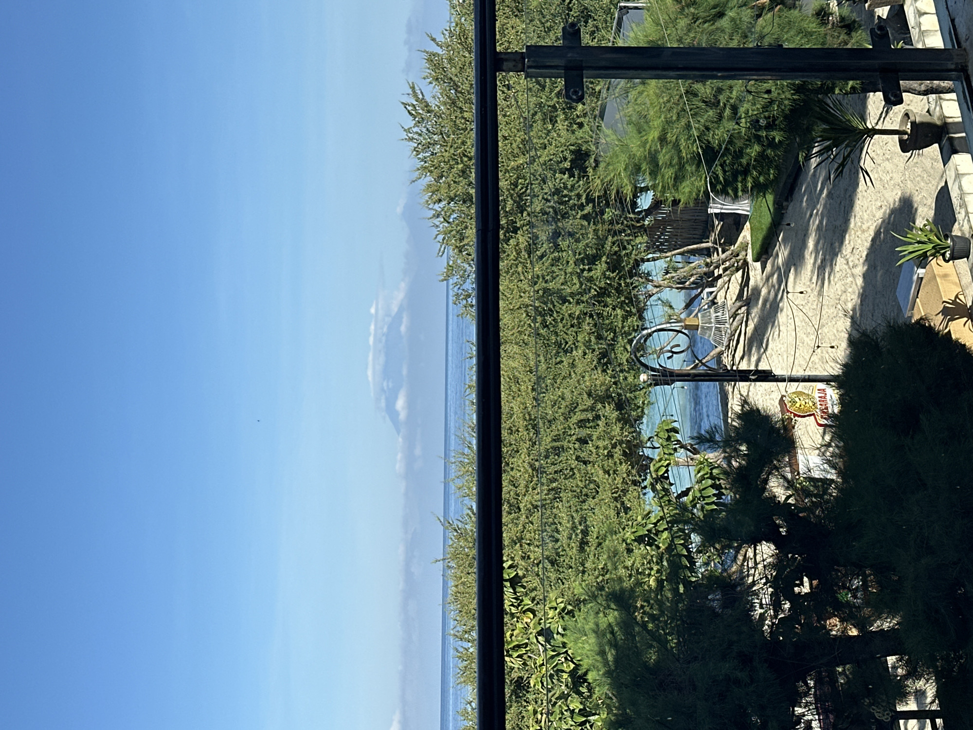 The image presents a view from an elevated position, possibly a balcony or terrace, in an area near the coast. The sky is a clear blue, transitioning to the darker blue of the ocean. A black metal frame is centrally positioned, possibly part of the balcony structure. Dense green foliage fills the mid-ground, obscuring some of the view. In the right portion of the image, hints of a patio or outdoor seating area are visible.
The location is in the city of Unknown, Indonesia. There are no people visible.