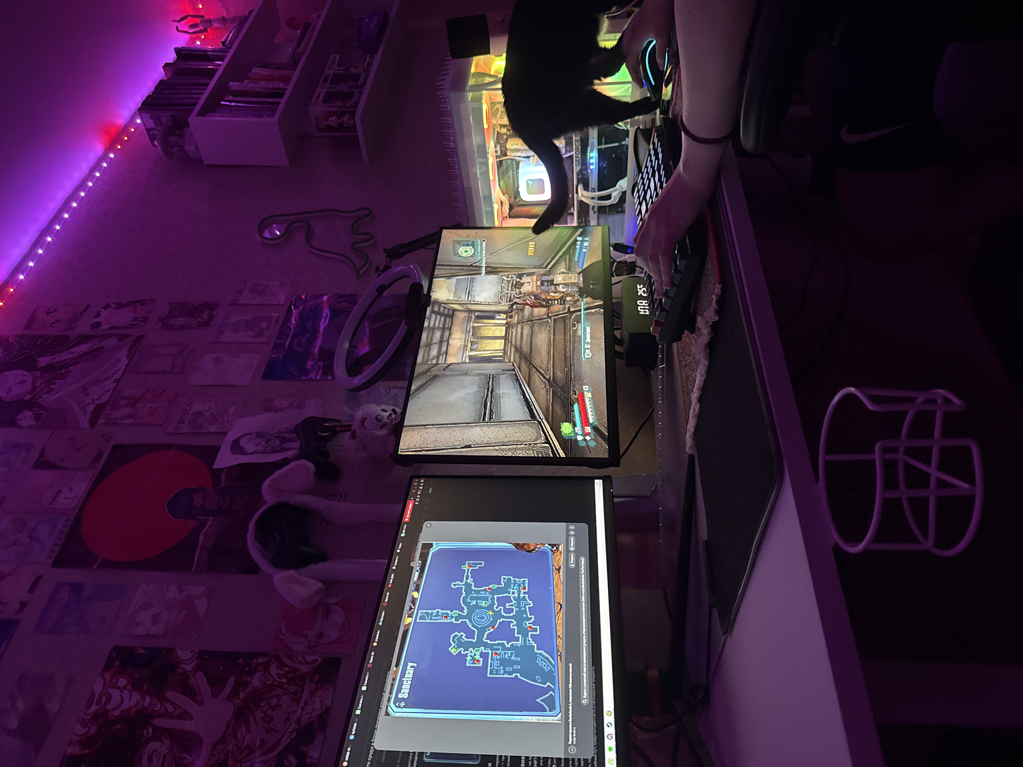 The image depicts a dimly lit room, likely a home office or gaming setup, with a strong purple ambient light source, possibly from LED strip lights. Two computer monitors are prominent. The left monitor displays a map with the title "Sanctuary." The right monitor shows a first-person perspective of a video game, featuring a character in a hallway-like environment. A black cat is walking across the desk, its tail extended. A person's hands are visible, operating a keyboard and a gaming mouse. The desk also holds a small digital clock displaying "BNA 25°". In the background, shelves are filled with books and various figurines. The walls are decorated with posters and artwork. A ring light is mounted on a stand behind the left monitor. A cup holder is attached to the side of the desk. The overall atmosphere is that of a dedicated gaming space, illuminated by colored lights, with a cat present. There are no discernible landmarks or features that specifically indicate a location within Pudomyagi, Russia.