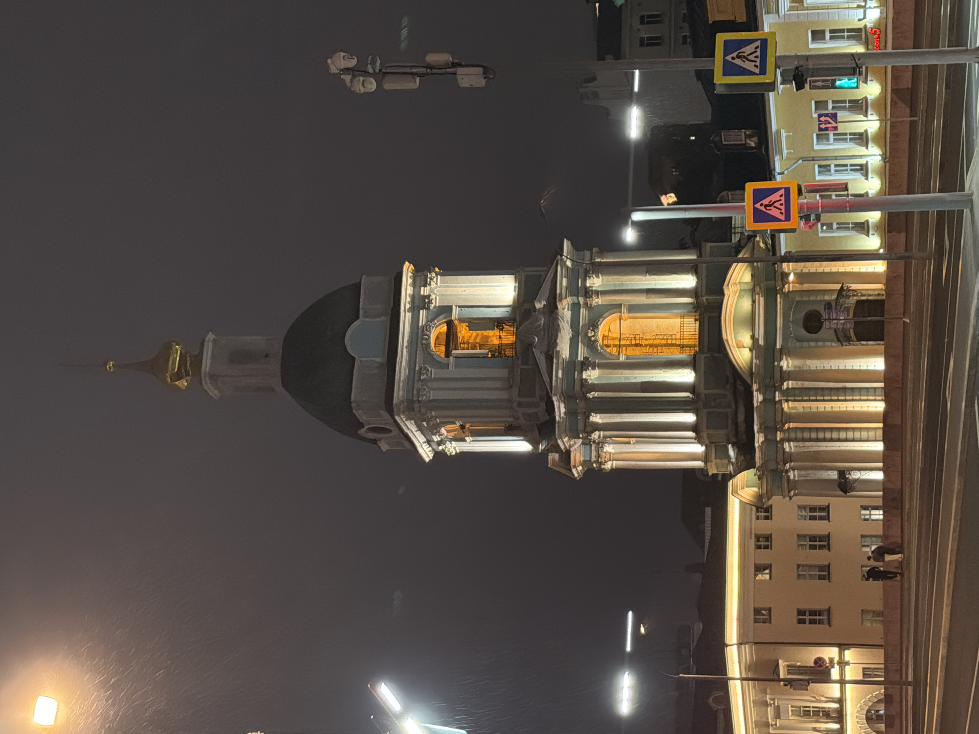 The image shows St. Nicholas Naval Cathedral in Moscow, Russia, at night. The cathedral's illuminated facade is visible. In the foreground, there are pedestrian crossing signs and lights, indicating a street scene. The environment appears to be a city street with buildings in the background. There are a few people near the base of the building.