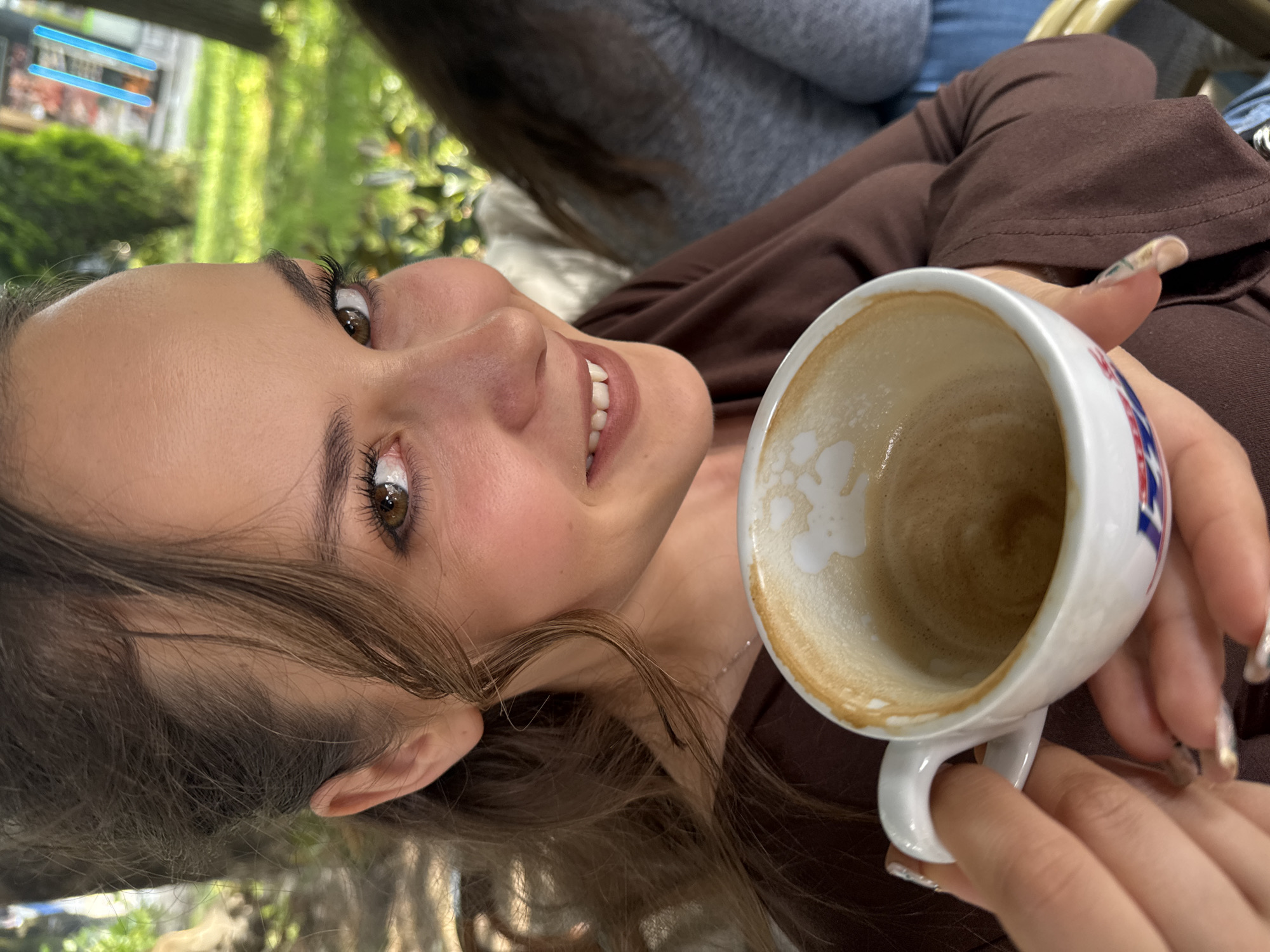 A woman with brown hair and eyes is sitting at an outdoor cafe, holding a white mug with a small amount of liquid inside. She is wearing a brown long-sleeved shirt. In the background, there is another person wearing a grey shirt and blue jeans. The location appears to be in a city, with greenery visible. The city is not explicitly identified.