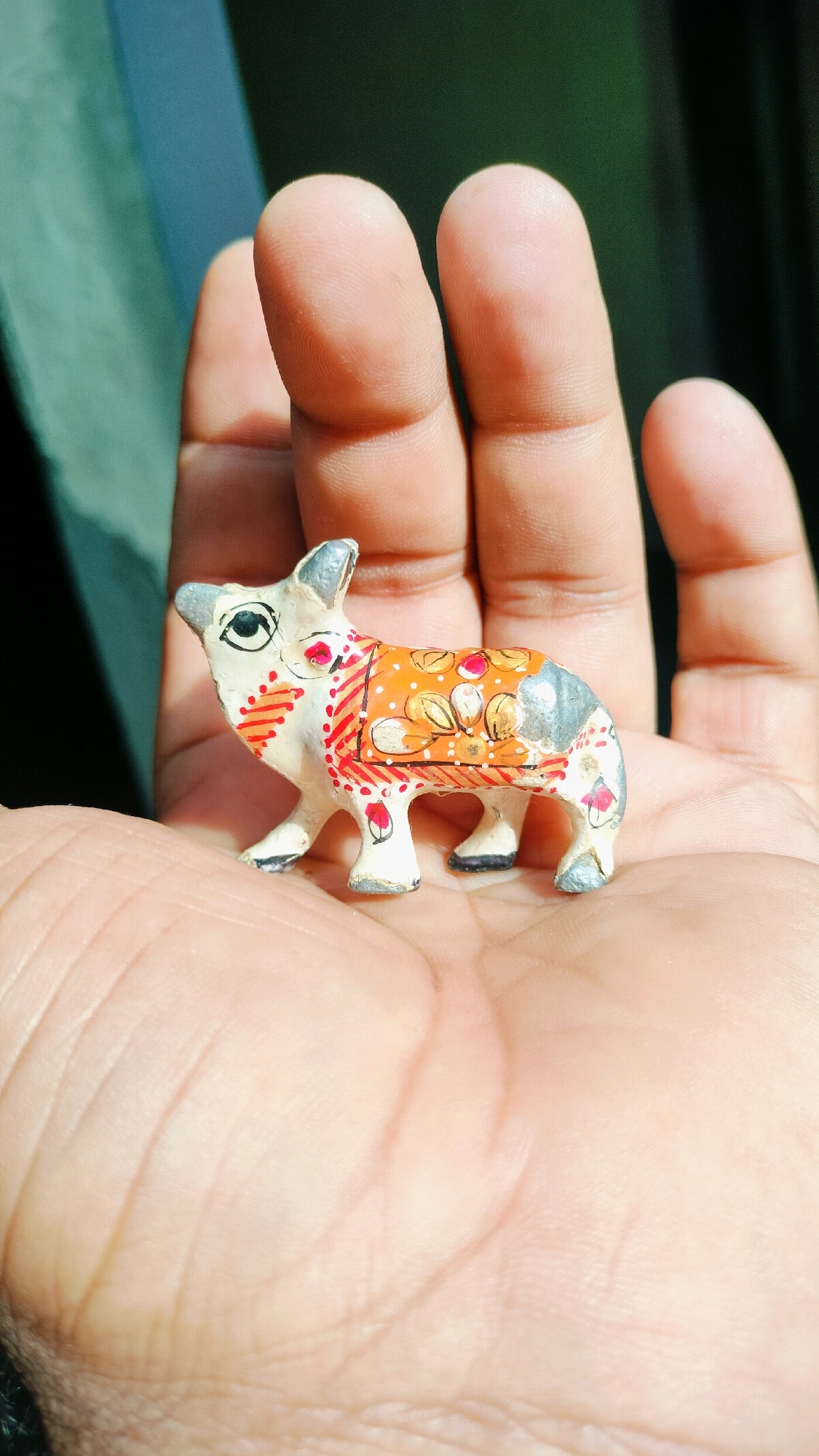 A macro shot shows a small, hand-painted figurine of a cow resting in the palm of a person's hand. The cow is predominantly white with gray accents on its ears and hindquarters. Its body is adorned with an orange saddle featuring floral designs in gold and red. Red and pink dots are painted along its neck and legs, and it has black eyes. The hand, with visible palm lines and fingerprints, supports the figurine, which is positioned facing left. The background is blurred, with a dark green area above and to the left, suggesting an outdoor setting. The lighting suggests the image was taken in daylight.