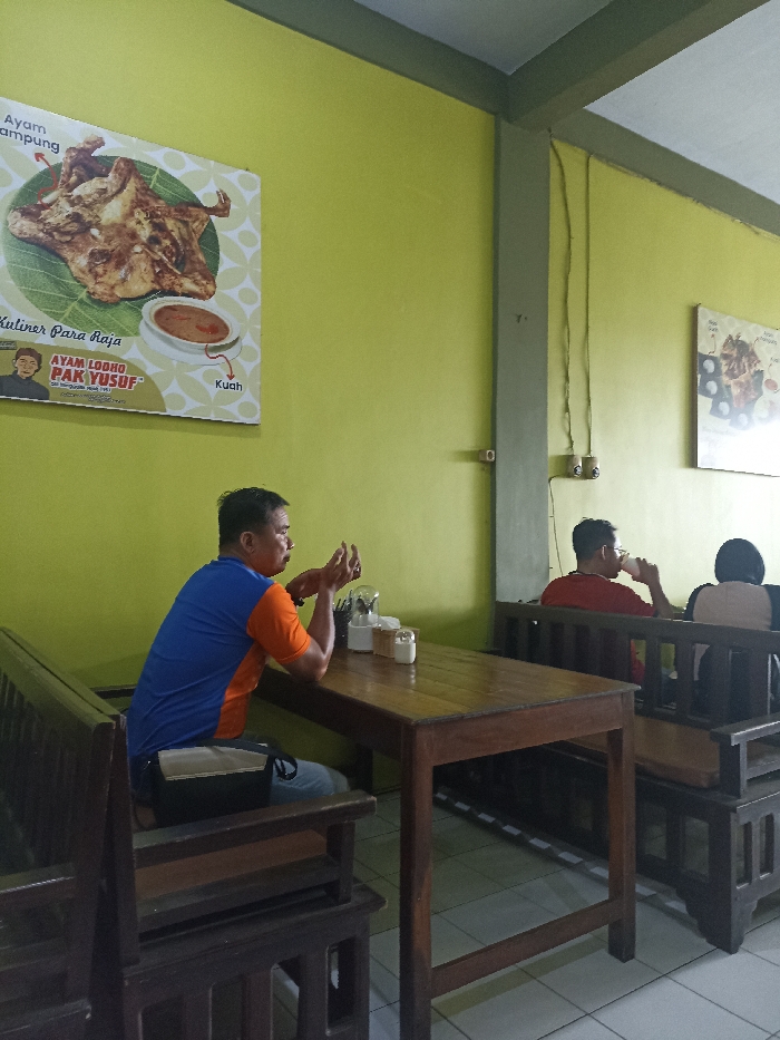 The image captures the interior of a casual eatery or restaurant, likely in Tulungagung, Indonesia, given the context and visible text. The walls are painted a vibrant lime green, contrasting with the dark wooden tables and benches that furnish the space. Grey support beams and visible electrical wiring run along the walls and ceiling. The floor is tiled.
In the foreground on the left, a man is seated at a wooden table. He wears a two-toned blue and orange short-sleeved shirt and light-colored trousers, with a black and beige bag resting on his lap. He is seen in profile, gesturing with his hands as if engaged in conversation or deep in thought, looking towards the right side of the frame. On his table are various condiments in small containers, including salt, sugar, and what appears to be a tissue holder.
Further back on the right, two other patrons are seated at another wooden bench. One man, wearing a red shirt and glasses, is visible from the back and side, appearing to drink from a cup. Next to him, another person, possibly a woman, is seen from behind, wearing a light-colored top and a dark head covering, suggesting a hijab. They appear to be relaxing or enjoying their meal.
A prominent poster hangs on the left wall, advertising a dish called "Ayam Lodho Pak Yusuf." It features an image of a whole grilled chicken served on a banana leaf, accompanied by a small bowl of sauce labeled "Kuah" (broth/sauce). The text on the poster also reads "Kuliner Para Raja" (Cuisine for Kings) and "Ayam Kampung" (Free-range chicken), along with a cartoon depiction of "Pak Yusuf." A second, partially visible poster on the right wall displays other food items. The overall atmosphere seems relaxed and informal, suggesting a daytime setting indoors.