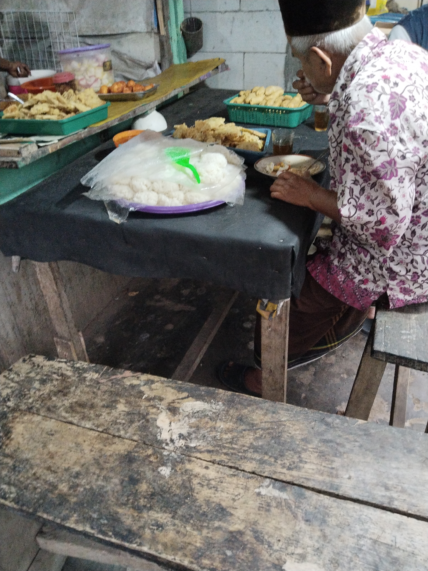 An overhead, eye-level shot captures a man in a floral shirt and brown pants sitting at a rustic wooden table in what appears to be an outdoor food stall in Cemplang, Indonesia. The man, with graying hair and wearing a dark cap, is actively eating, his hands near his mouth. A plate of food is in front of him, along with a glass of liquid.
The table is covered with a dark cloth, on which rests a large plastic bag filled with white rice. To the left, a green plastic container holds a pile of fried food, and another similar container is visible further back, also filled with fried items. A yellow shelf runs along the back, displaying more containers of food and some scattered utensils. The background shows a rough concrete wall and metal shelving, suggesting a simple, functional establishment. In the foreground, a weathered wooden bench runs across the bottom of the frame, adding to the rustic ambiance of the scene. The lighting is somewhat dim, with a natural light source, possibly from the side, illuminating the food and the man.