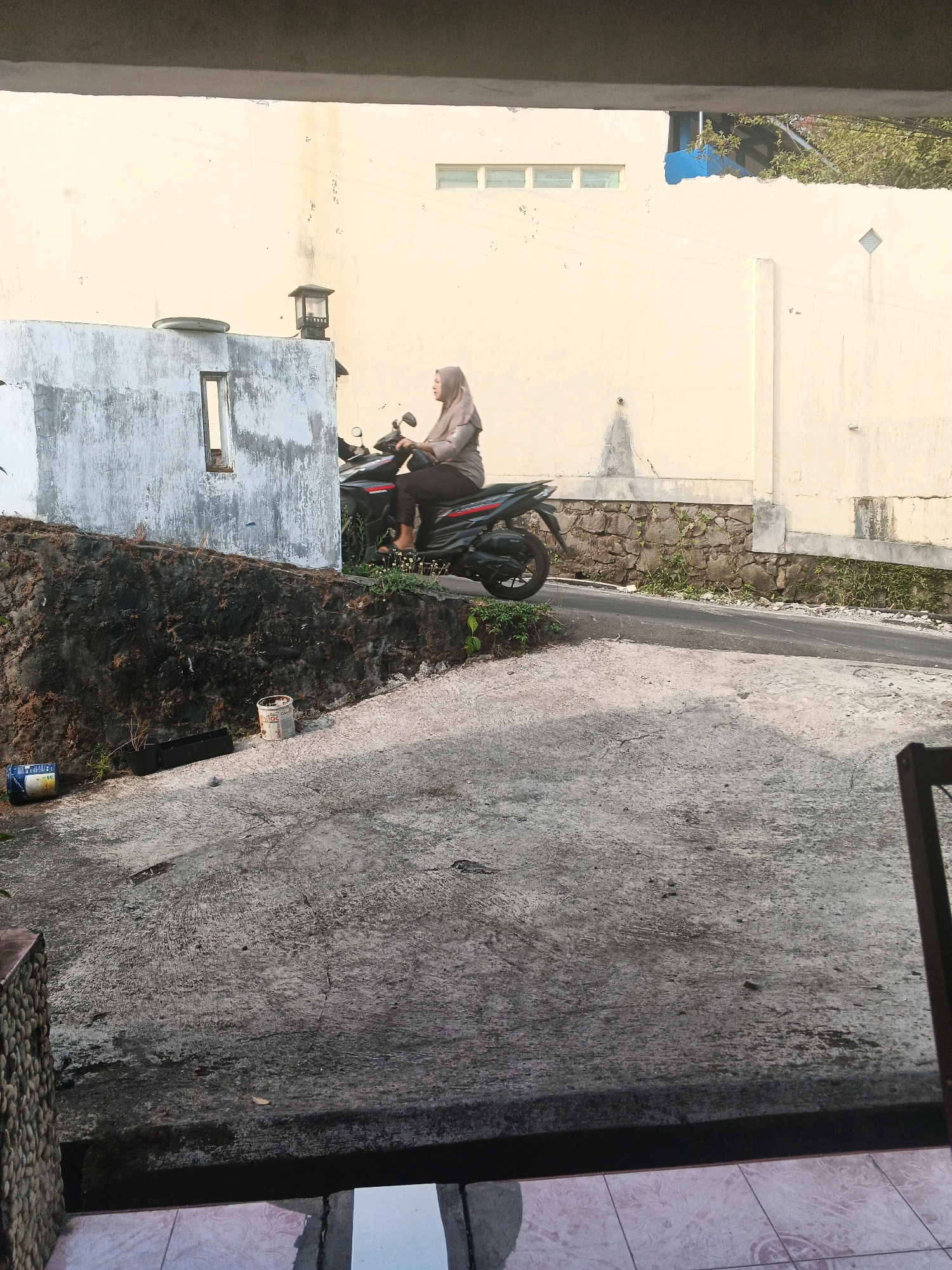 A woman wearing a headscarf and a light-colored top and dark pants sits astride a black motorcycle. She is facing left and appears to be stationary on a paved road that slopes uphill. Behind her, a textured, light-colored wall extends horizontally across the scene. To the left of the motorcycle, there is a white, rough-textured wall with a rectangular opening. The ground in the foreground is a rough, gray concrete surface. A stone wall runs along the left side of the road. There are two cylindrical containers and a black rectangular planter on the ground near the stone wall. The location is identified as Keji, Indonesia.
