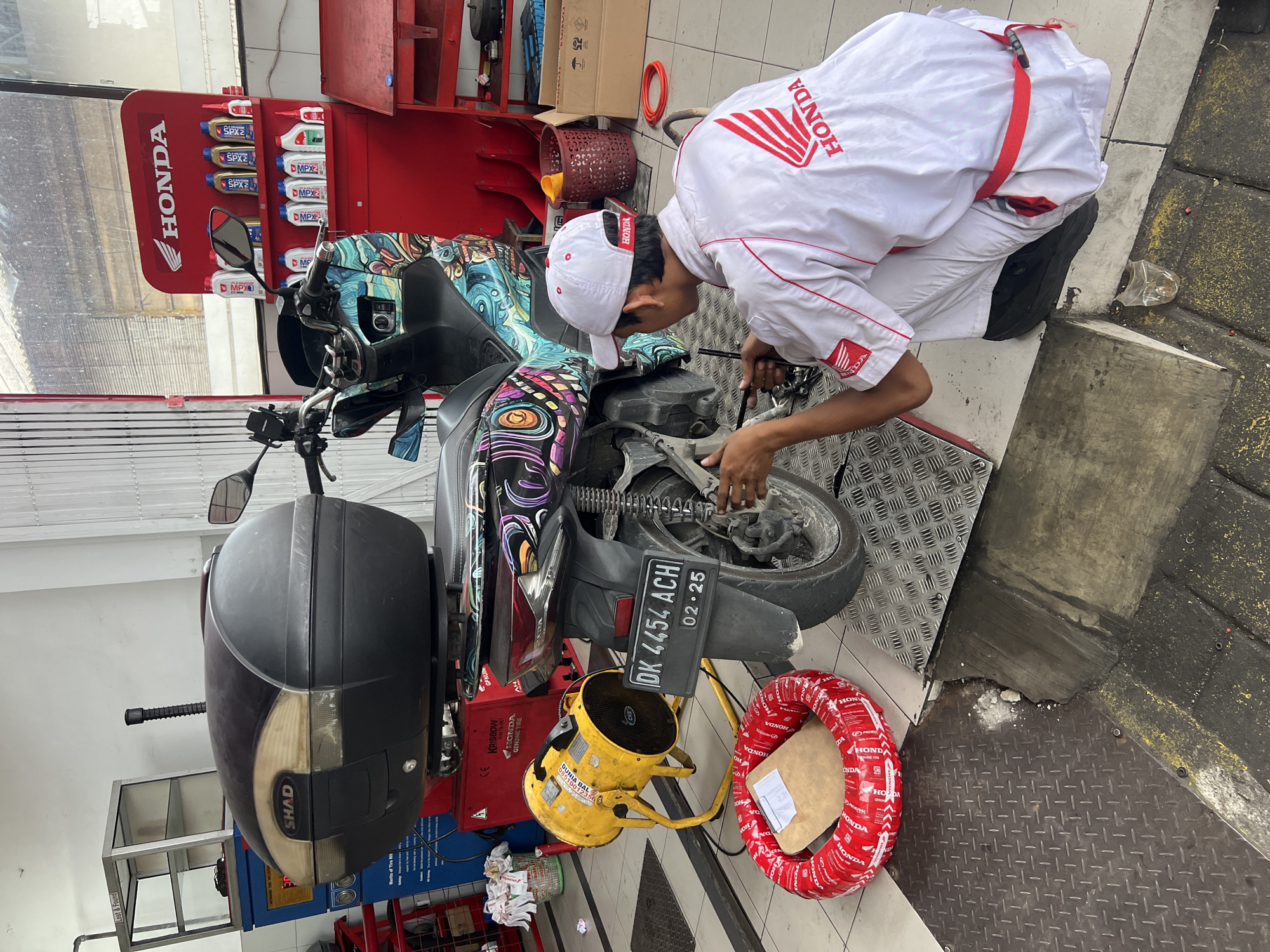 The image captures a detailed scene inside a motorcycle service or repair shop, likely an authorized Honda center, located in Kuta Selatan, Indonesia. A male mechanic, dressed in a white Honda uniform (shirt and cap with the Honda logo) and light-colored pants with a red shoulder strap, is kneeling on the checkered metal floor. He is actively engaged in servicing the rear wheel of a scooter, holding a tool with focused attention.
The motorcycle itself is distinctive, featuring a vibrant, multi-colored abstract design covering its body. A black SHAD brand storage box is mounted on the rear. Its Indonesian license plate, "DK 4454 ACH," is clearly visible, along with an expiration date of "02-25" (February 2025), confirming the Bali/Nusa Tenggara Barat region.
The shop environment includes a red display stand prominently featuring Honda motor oils such as SPX1, SPX2, MPX1, and MPX2. Various workshop equipment is present, including a yellow industrial fan labeled "DUNIA BAL" with a partial phone number (08510...). A new red Honda genuine tire, wrapped in plastic with "PEDOMAN SNI" (Standard Nasional Indonesia) branding, rests on the floor. Other visible text includes "Lost & Found" on a cabinet, "KRISBOW KW19-967 HONDA GENUINE TIRE" on a red machine, and "Merits of Tire Nit" on another piece of equipment. The overall setting indicates a professional and well-equipped service bay.