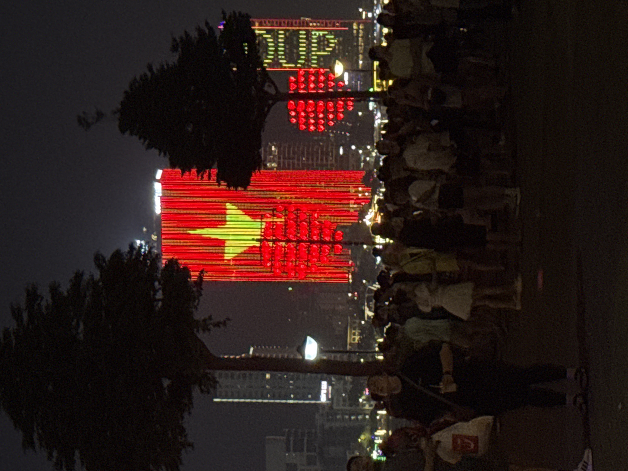 At night, a crowd of people stand on a walkway. A building in the background displays a red and yellow flag, resembling the flag of Vietnam, with a prominent yellow star. Further back, another building is illuminated with the word "DUP" and a heart shape made of red dots. Trees are silhouetted in the foreground. A street lamp is visible in the midground. This scene is likely in Đà Nẵng, Vietnam.