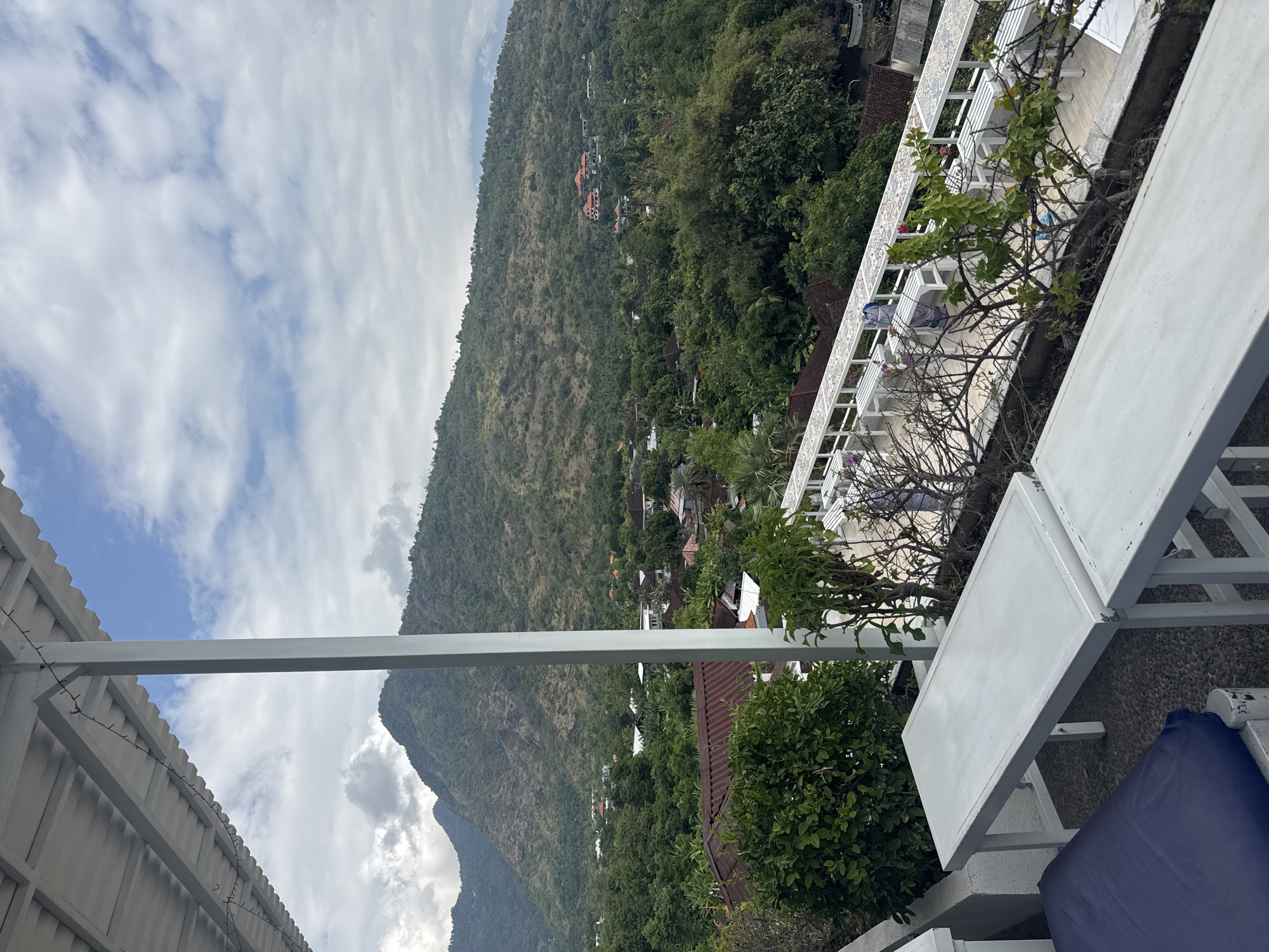 The image shows a view from an elevated outdoor seating area, likely a cafe or restaurant, overlooking a lush green valley with mountains in the background. In the foreground, there are white tables and benches, some with blue cushions. A white support beam and roof structure are visible on the left and top of the frame. Beyond the seating area, a village or town is nestled amongst dense tropical foliage, with red-tiled roofs and white buildings dotting the landscape. The mountains in the distance are covered in greenery and appear somewhat dry in places. The sky is partly cloudy with patches of blue. The scene suggests a scenic location in a rural or semi-rural area, potentially within the Abang district of Bali, Indonesia, known for its natural beauty and hillside villages. No people are actively engaged in any interactions within the frame.