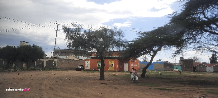 A street scene shows a dirt road with various structures visible. An orange building with posters is prominent, along with smaller shacks and houses. Trees provide shade, while electrical poles and wires traverse the sky. A motorcycle is seen on the right, and a car is parked near the orange building. The sky is partly cloudy, with a mix of gray and blue.
