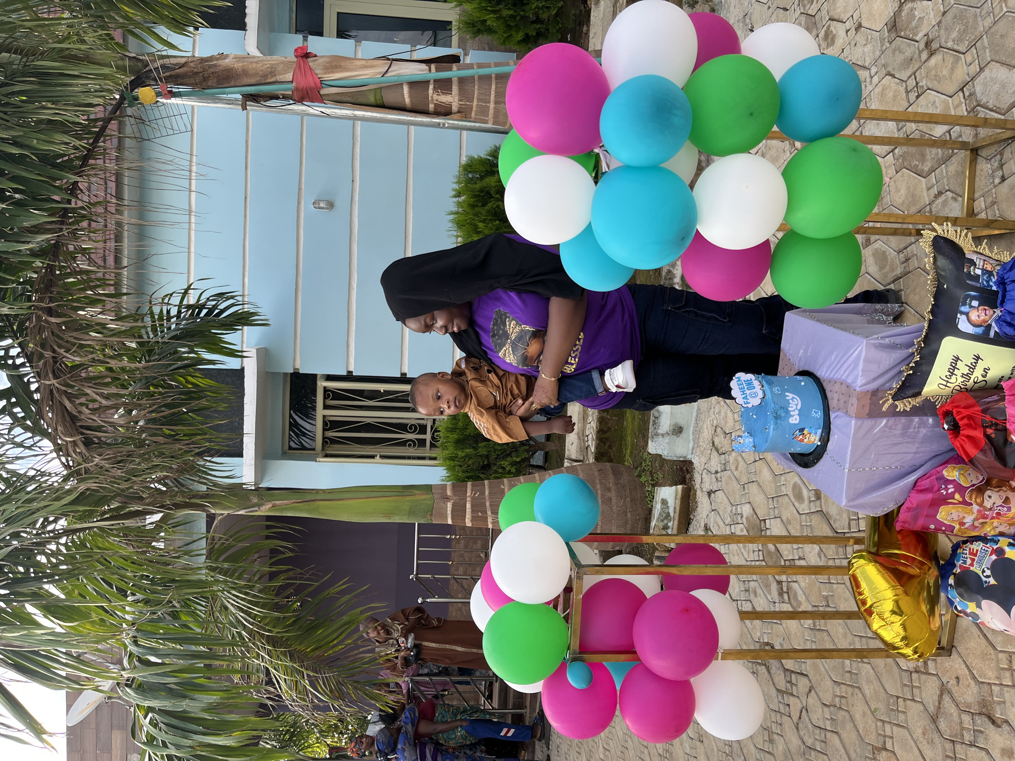 The scene depicts an outdoor birthday celebration in Jos, Nigeria, featuring multiple individuals and festive decorations.
In the foreground, a light-colored, interlocking paver stone pathway serves as the ground. Two gold-colored, rectangular metal stands display clusters of balloons in solid colors: magenta, white, green, and cyan. Between these stands, a small table covered with a light purple fabric holds a round, two-tiered blue birthday cake decorated with characters from the "Bluey" television show and a small banner reading "FAHEEM @ ONE." Nearby on the ground are various party items, including a gold foil balloon shaped like the numeral "1," and several colorful drawstring bags, one featuring Disney Princesses and another Mickey Mouse characters. A black picture frame with gold filigree, containing a photograph of a child and text that reads "Happy Birthday Son," is also visible.
The central figures include a woman with dark skin, appearing to be in her late 20s to early 30s, wearing a black hijab, a purple t-shirt with a graphic of a woman's face and the word "BLESSED," and dark pants. She holds a male infant, approximately one year old, dressed in a brown long-sleeved shirt, blue jeans, and white sneakers. The infant's gaze is directed towards the right side of the frame. The woman is looking left with a slight smile.
To the left of the central figures, a group of individuals is partially visible. One woman in a blue patterned outfit holds a child. Another woman, dressed in a brown traditional garment, stands by a dark metal railing, looking down at an object in her hands.
The background features several large palm trees with green fronds, some exhibiting signs of dryness. A light blue, multi-story residential building with white horizontal trim bands is prominent on the right, featuring windows secured with white security grates. Lush green shrubbery borders the pathway in front of this building. On the left, a darker-colored building, possibly brick or paneled, is partially visible, with a white satellite dish mounted above. The sky is light and somewhat hazy.