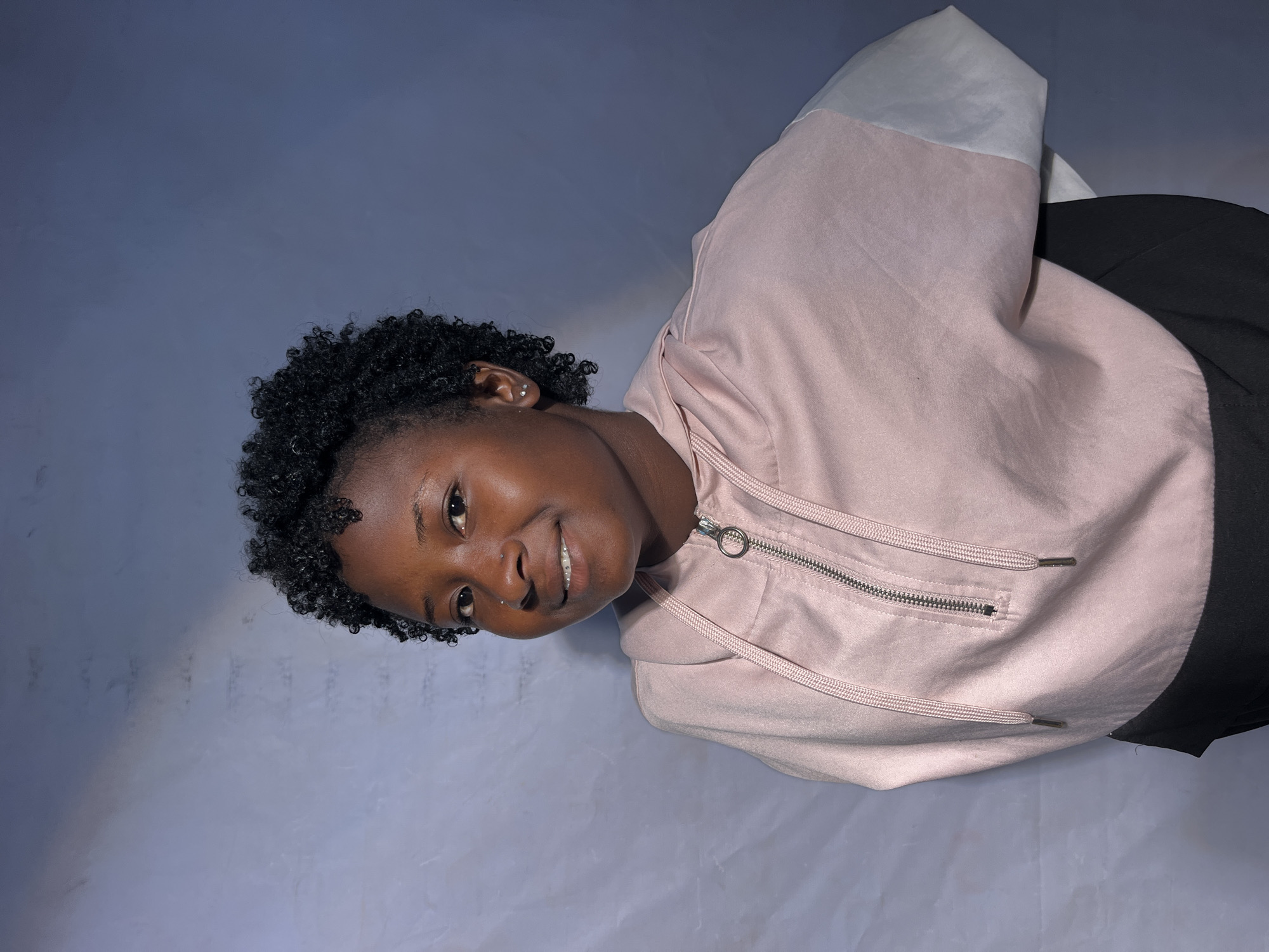 A young woman with short, curly dark hair is pictured from the chest up, looking to her left with a slight smile. She has a small nose stud and appears to be wearing braces. She is wearing a light pink hooded jacket with a zipper and drawstrings. The background is a textured, mottled grey and blue fabric. The image is a portrait.