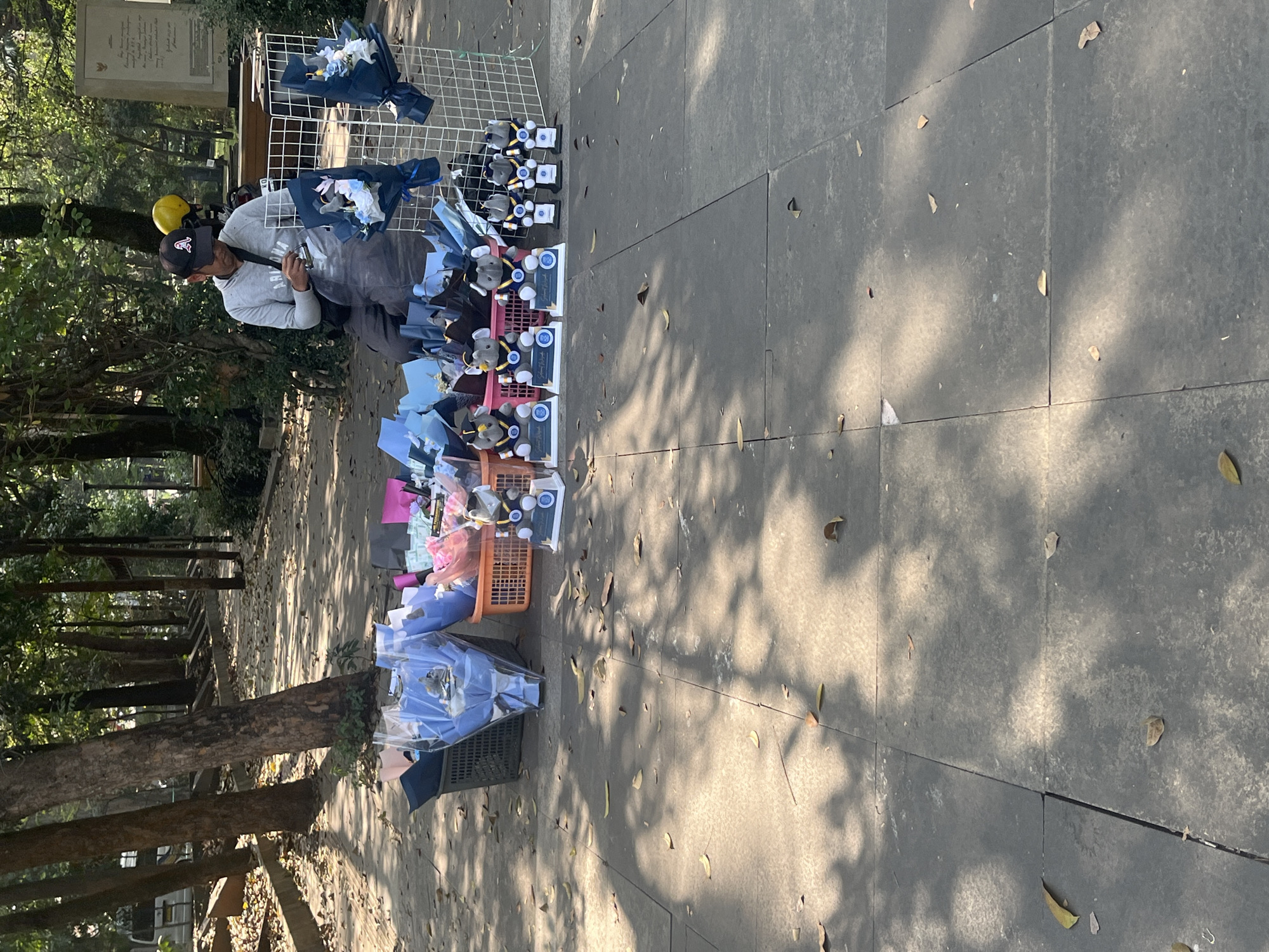 The image depicts a street vendor selling bouquets and stuffed animals in a park-like setting. The vendor, a man wearing a gray hoodie and a baseball cap, is sitting on the ground, attending to his merchandise. The bouquets are wrapped in plastic and some are displayed in baskets, while others are placed on a white wire rack. The stuffed animals appear to be elephants wearing graduation caps and sashes. The ground is paved with square tiles, and fallen leaves are scattered across the surface. Sunlight filters through the trees, creating dappled shadows on the ground. In the background, trees and a stone monument are visible, suggesting an outdoor public space. The scene is likely in Bandung City, Indonesia, given the presence of street vendors selling such items in public parks, which is a common practice there.