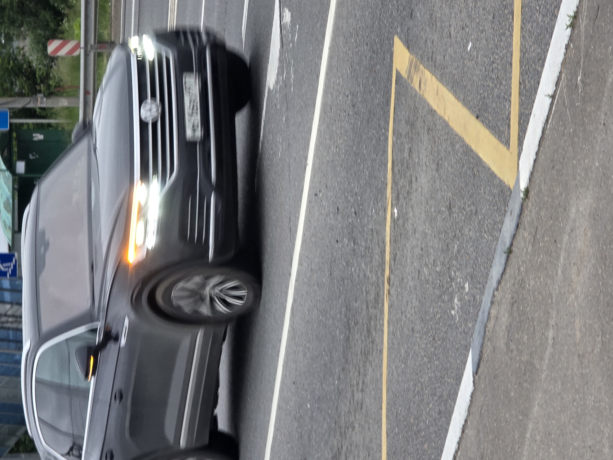 The image shows a dark-colored SUV driving on a paved road. The car is angled, with its front end facing the viewer. The headlights are illuminated, and the turn signal on the passenger side is also lit, indicating the vehicle is in motion and potentially turning or changing lanes. The car's license plate is partially visible and appears to have Russian characters. The road has yellow and white lane markings, suggesting it's a public street or highway. The background includes a metal guardrail, some greenery, and a red and white striped pole, which could be a marker or part of a safety barrier. The lighting suggests it could be daytime, possibly overcast, given the lack of strong shadows. The scene appears to be a typical urban or suburban environment. There is a blue sign with a white wheelchair symbol visible in the background to the left, indicating a disabled parking area or accessibility feature. The overall impression is a candid shot of a vehicle in transit.