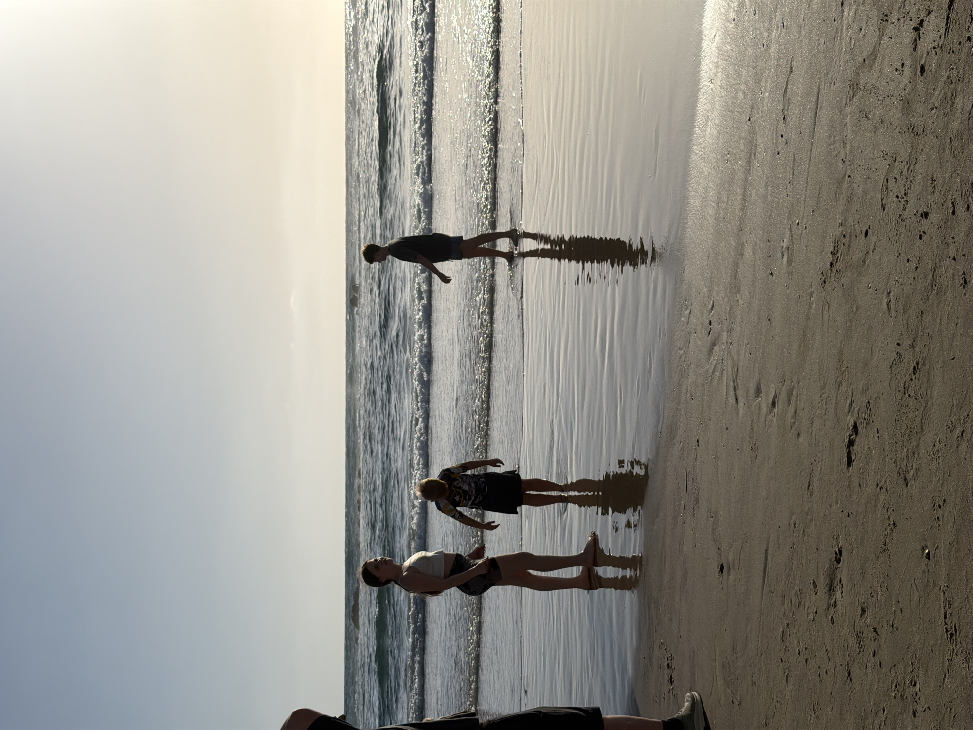The image shows a beach scene in Bat Yam, Israel. Three people are standing on the wet sand near the water's edge. One person is closer to the water, and two others are positioned slightly further back. The scene appears to be during sunset or sunrise, with the light casting long shadows. The beach is sandy, with visible footprints. The water is wavy.