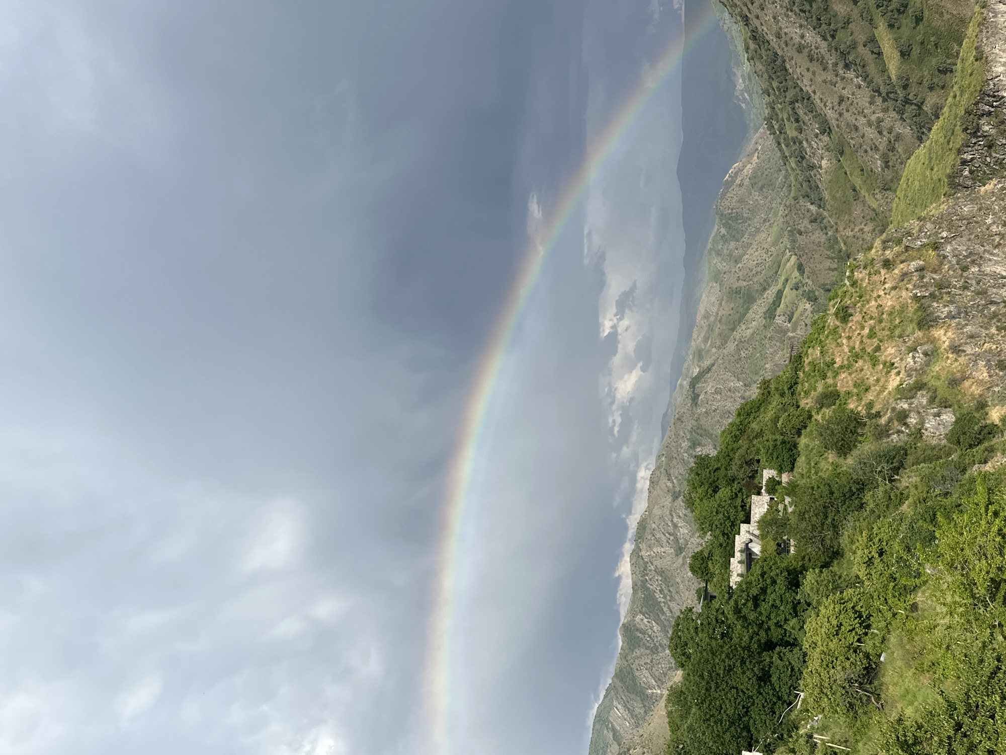 The image depicts a dramatic landscape in Garni, Armenia, featuring a vibrant rainbow arcing across a cloudy sky over a rugged, mountainous terrain. The foreground is dominated by lush green trees and dense foliage clinging to steep slopes, with hints of dry grass and rock formations. A series of stone structures, possibly buildings or ruins, are nestled amongst the trees on the hillside. The middle ground reveals the undulating contours of the mountains, characterized by a patchwork of green vegetation and rocky outcrops, with deep valleys carved between them. The sky above is a mix of dark, heavy clouds and lighter patches, with the rainbow serving as a striking contrast. The overall scene suggests a post-rain atmosphere in a natural, somewhat wild environment. There are no people visible in the image.