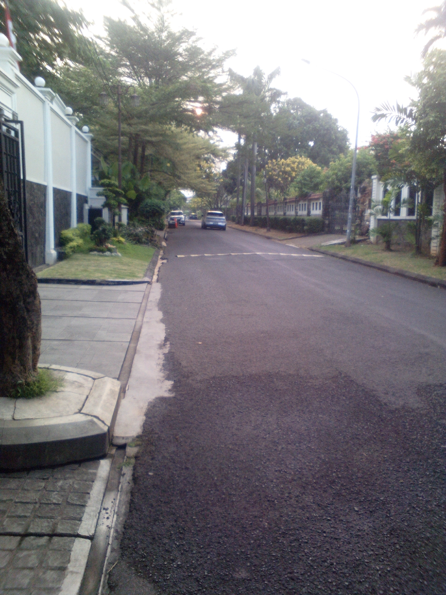 A residential street in South Jakarta, Indonesia, is depicted with a paved asphalt road on the right and a sidewalk made of square concrete tiles on the left. A white, gated wall with dark stone accents runs along the left side of the street, bordering a well-maintained lawn with manicured bushes and small trees. A large tree trunk is visible in the foreground on the left. Several cars, including a blue sedan and a silver sedan, are parked or driving on the road, suggesting a quiet urban environment. The sky is overcast and bright, indicating daytime. The architecture of the houses visible in the background suggests a modern residential area.