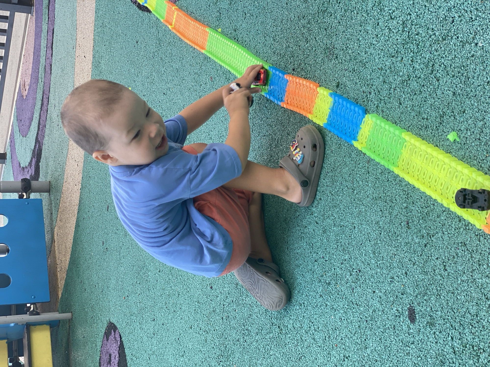 In Sochi, Russia, a young East Asian boy is kneeling on a bright green, textured playground surface. He is wearing a blue t-shirt and coral shorts, with grey Crocs on his feet. The boy is actively playing with a colorful toy car track, made of interconnected green, blue, and orange segments. He is holding a small red car and a white and black object, possibly another toy car or accessory, on the track. The playground equipment, including blue and yellow structures, is visible in the background. The ground is a uniform green with scattered small dark specks.