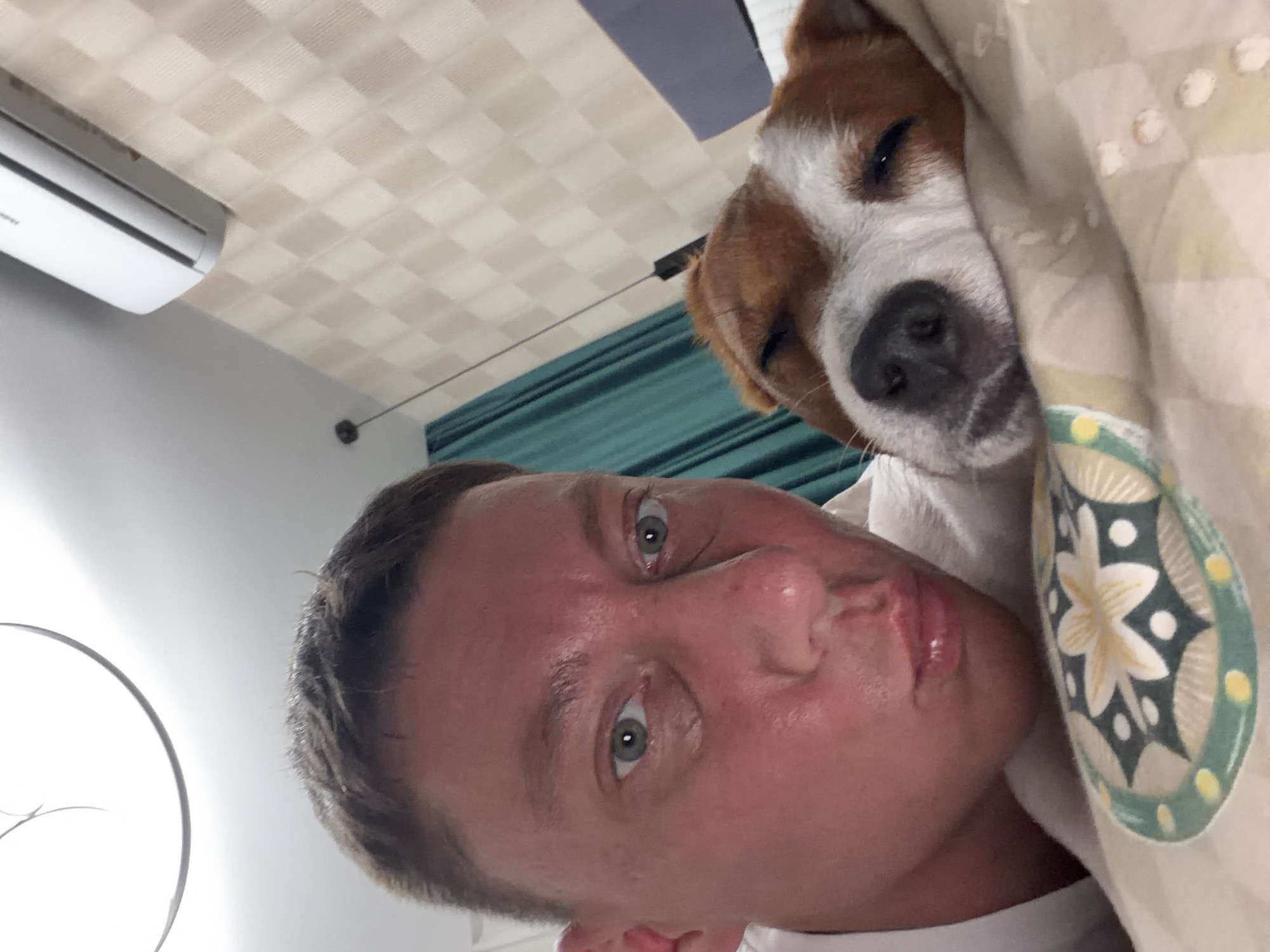 The image shows a close-up selfie of a person and a dog. The person is in the foreground, with their face turned towards the camera. They have short, dark hair, green eyes, and appear to be making a kissing face. Their skin has a reddish hue, particularly on the cheeks and nose, suggesting flushing or a reaction. The person is wearing a white shirt.
Next to the person, a Jack Russell Terrier dog is resting its head on a pillow. The dog has brown and white markings and its eyes are partially closed, appearing sleepy. The dog is nestled into a pillow with a patterned cover featuring a floral design in green, yellow, and white.
The background shows an indoor setting. To the right, a beige, textured wallpaper is visible. Above the wallpaper, a white air conditioning unit is mounted on the wall. To the left, a teal-colored curtain can be seen. A ceiling light fixture, circular and white, is visible at the top of the image.
The overall scene suggests a relaxed, domestic moment, possibly in a bedroom. There is no indication of a specific location within the city of Moscow, Russia.