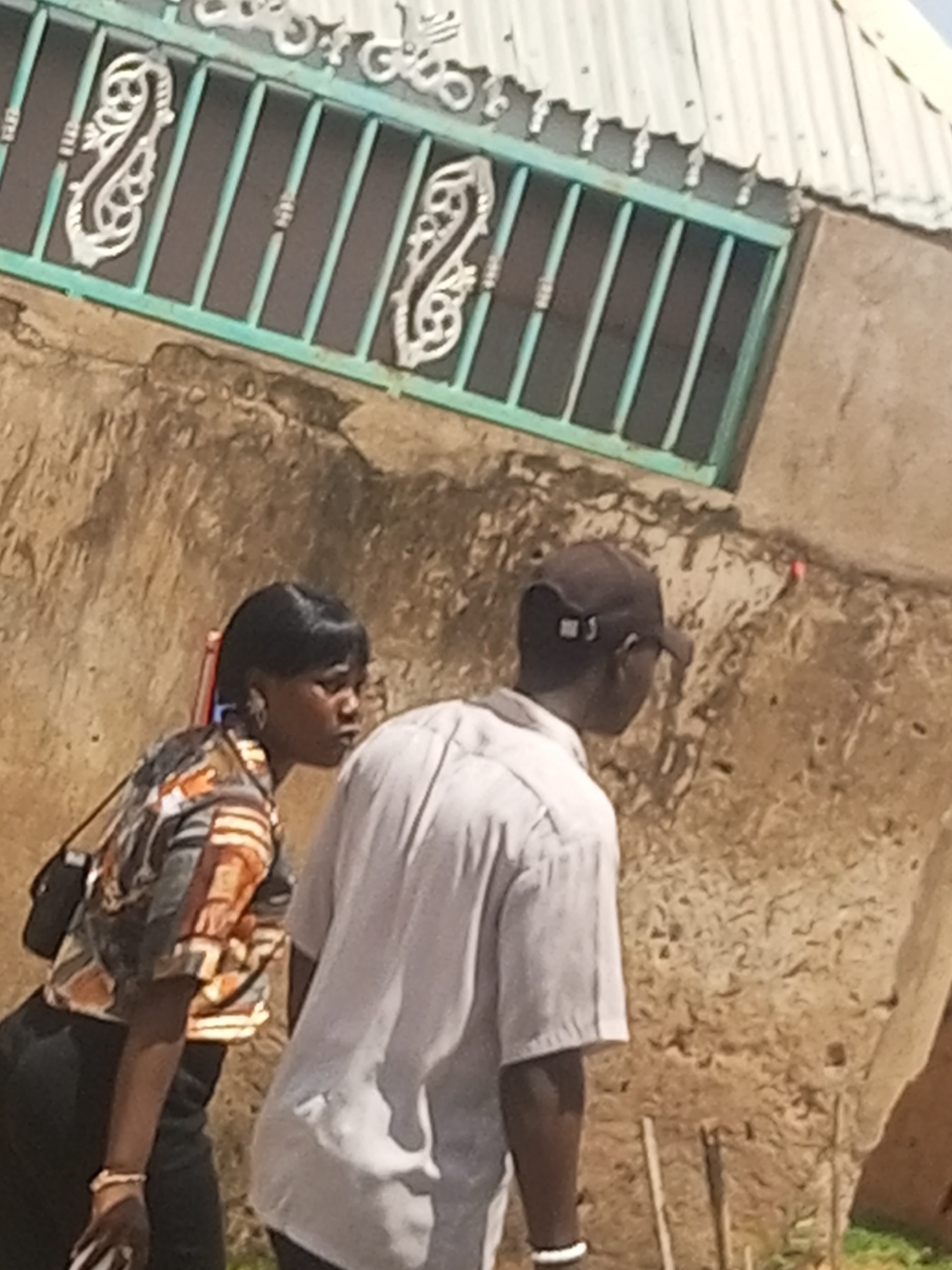 Two individuals, a woman and a man, are captured in profile, standing in front of a rustic, earth-toned wall. The woman, on the left, has dark hair pulled back and wears a patterned shirt with orange and brown hues, accessorized with a bracelet on her left wrist. A black shoulder bag rests on her right side. To her right, the man is wearing a light-colored collared shirt and a dark baseball cap. The background features a green metal gate with ornamental scrollwork, a corrugated metal roof above, and the textured mud wall of a building. The scene suggests an outdoor setting in a built environment.