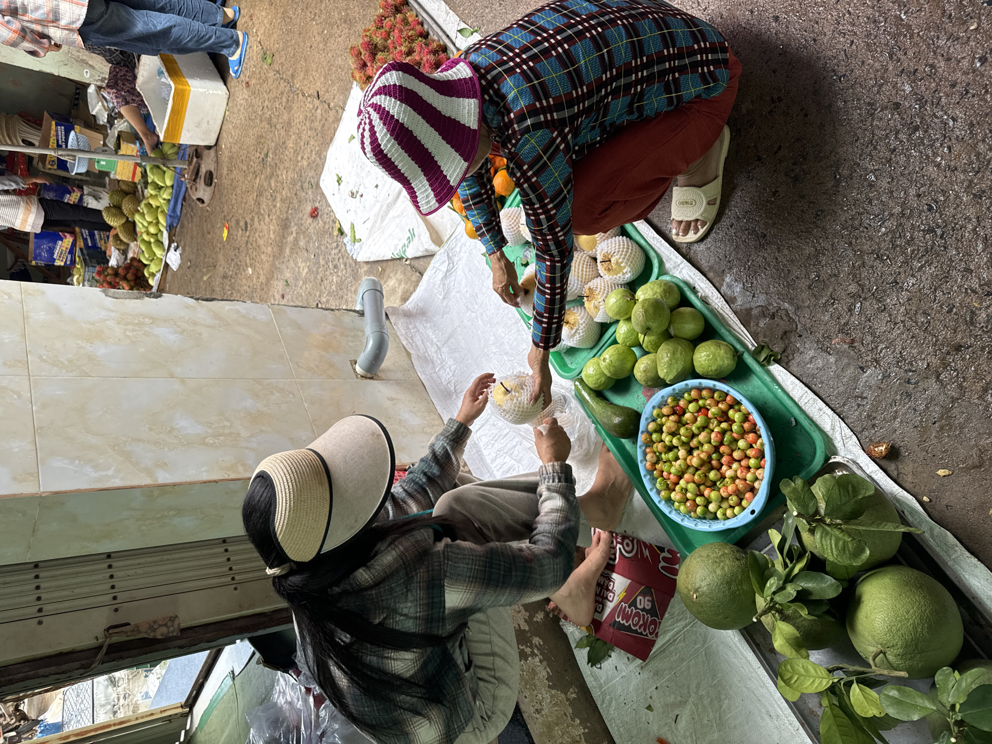 In Mũi Né, Vietnam, two individuals are engaged in sorting and packaging fruit at an outdoor market stall. The foreground features a variety of produce, including large green citrus fruits, a bowl overflowing with small, colorful berries, and several guavas. One person, wearing a plaid shirt and a wide-brimmed visor, is crouched on the ground, inspecting a piece of fruit wrapped in white netting. The other person, also in a plaid shirt and a distinctive striped hat, is seated on the ground, reaching towards a green tray of guavas. The background shows more fruit stalls with piles of durian, rambutans, and oranges, along with shoppers and vendor merchandise. The ground is wet and appears to be concrete or asphalt, typical of an outdoor market environment. The scene captures a moment of commerce and food preparation within a bustling local market.