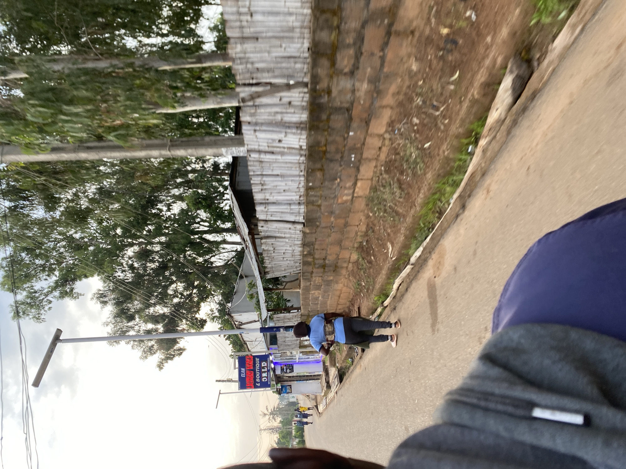 The image depicts a street scene in the city of Bar Jirgi Summa, Nigeria. In the foreground, a person's head and shoulder are visible, obscuring part of the view. A paved road runs through the scene, with a drainage ditch along the right side. A woman in a blue shirt and dark pants is walking away from the camera on the left side of the road. In the background, a shop with a sign that reads "ELAN LUXURY WARD & BOUTIQUE ORLD" is visible, with blue lighting emanating from within. Several other individuals, likely pedestrians, are also seen walking on the road in the distance. The environment includes tall trees and a bamboo fence. The sky is overcast with clouds.