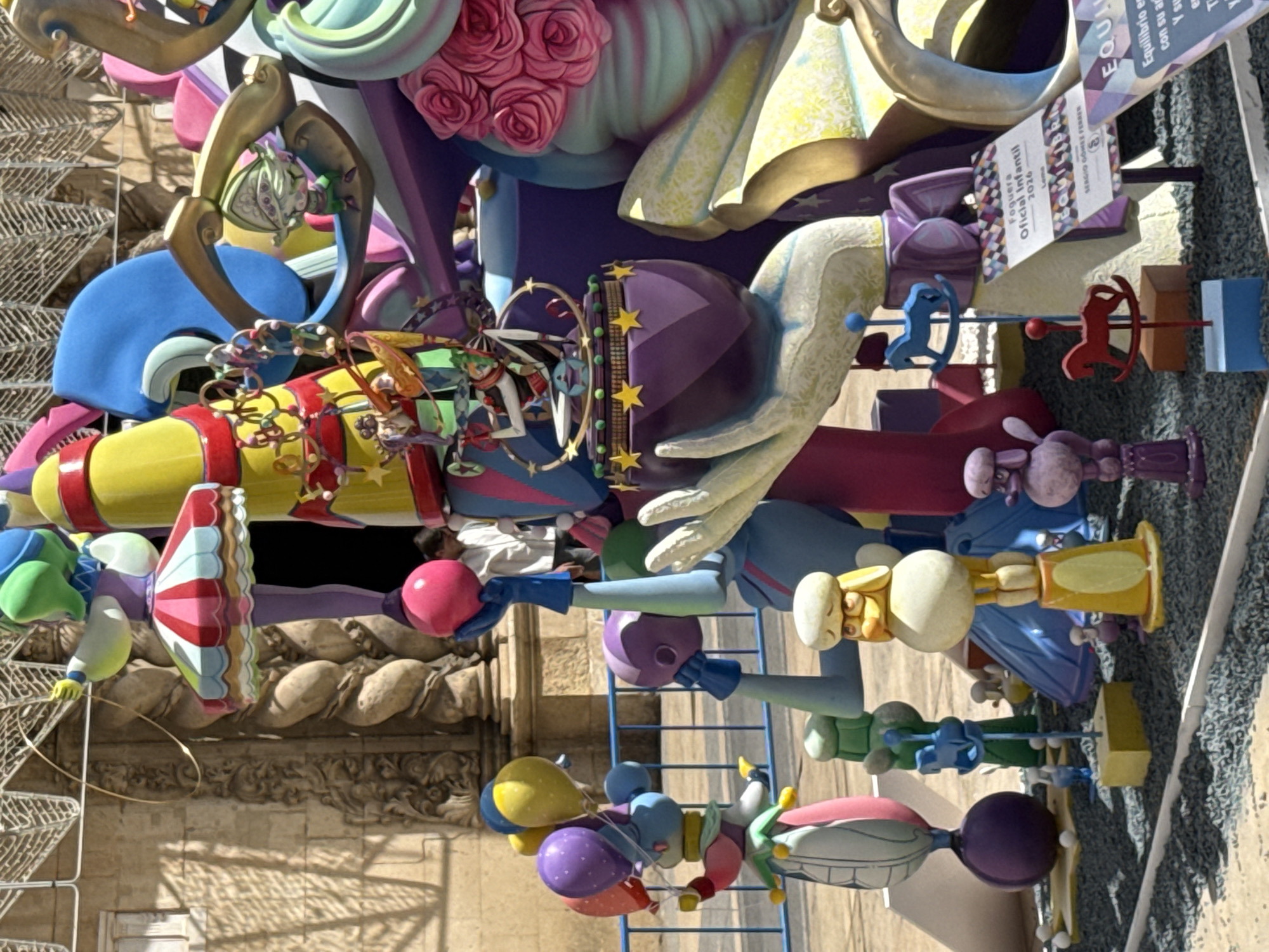 In the city of Unknown, Unknown, a colorful, elaborate sculpture stands in an outdoor setting. The sculpture is a whimsical, abstract creation with various figures and elements, including balloons, a ring-spinning character, and a large, stylized hand. The sculpture is constructed from bright, solid colors and appears to be made of a hard material, possibly fiberglass or painted wood. In the background, a stone building with ornate architectural details is visible, suggesting it might be a historic landmark. A blue metal fence runs across the middle ground, separating the sculpture from the viewer. Small, toy-like figures of horses and dogs are placed around the base of the sculpture, adding to the playful theme. A sign is visible on the right side of the sculpture, with text in Spanish. The ground is covered in dark gray gravel. The overall scene is vibrant and festive, likely a public art installation or part of a celebration.