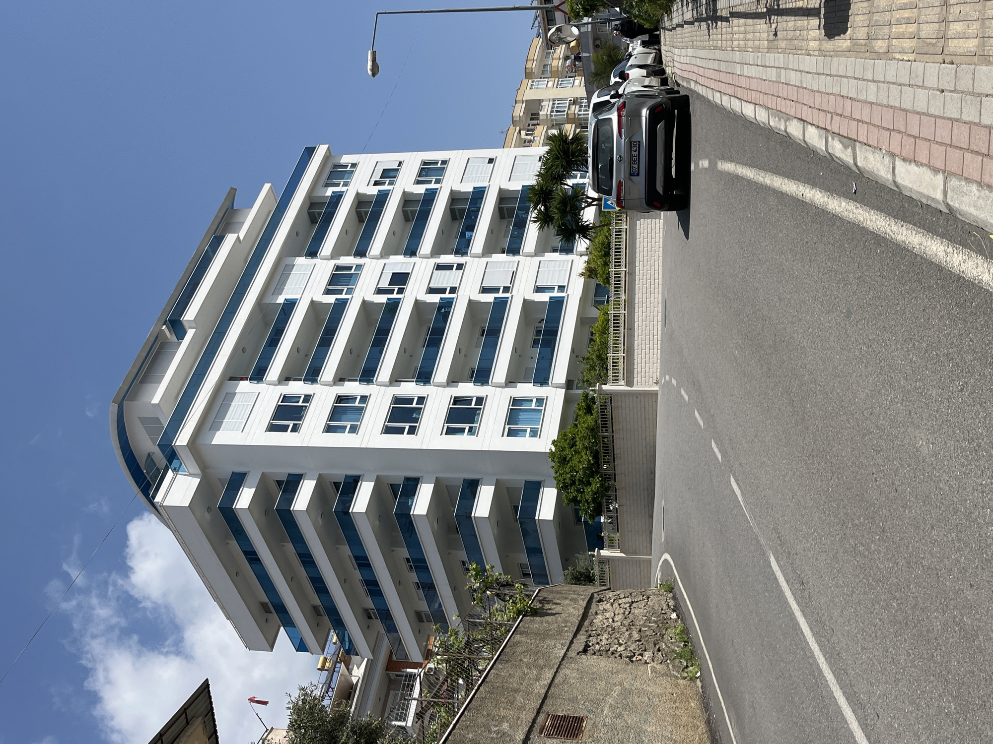 A daytime, ground-level shot, focuses on a modern, white building with blue-tinted balconies in Alanya, Turkey. The building rises to the left of the frame, with a rounded corner design on the upper floors. To the right of the building, a gray van is parked along the roadside. The street curves slightly to the right, marked with a white line. A sidewalk with brick and concrete segments runs along the right edge. In the background, other buildings are visible under a clear blue sky. The location is Alanya, Turkey.