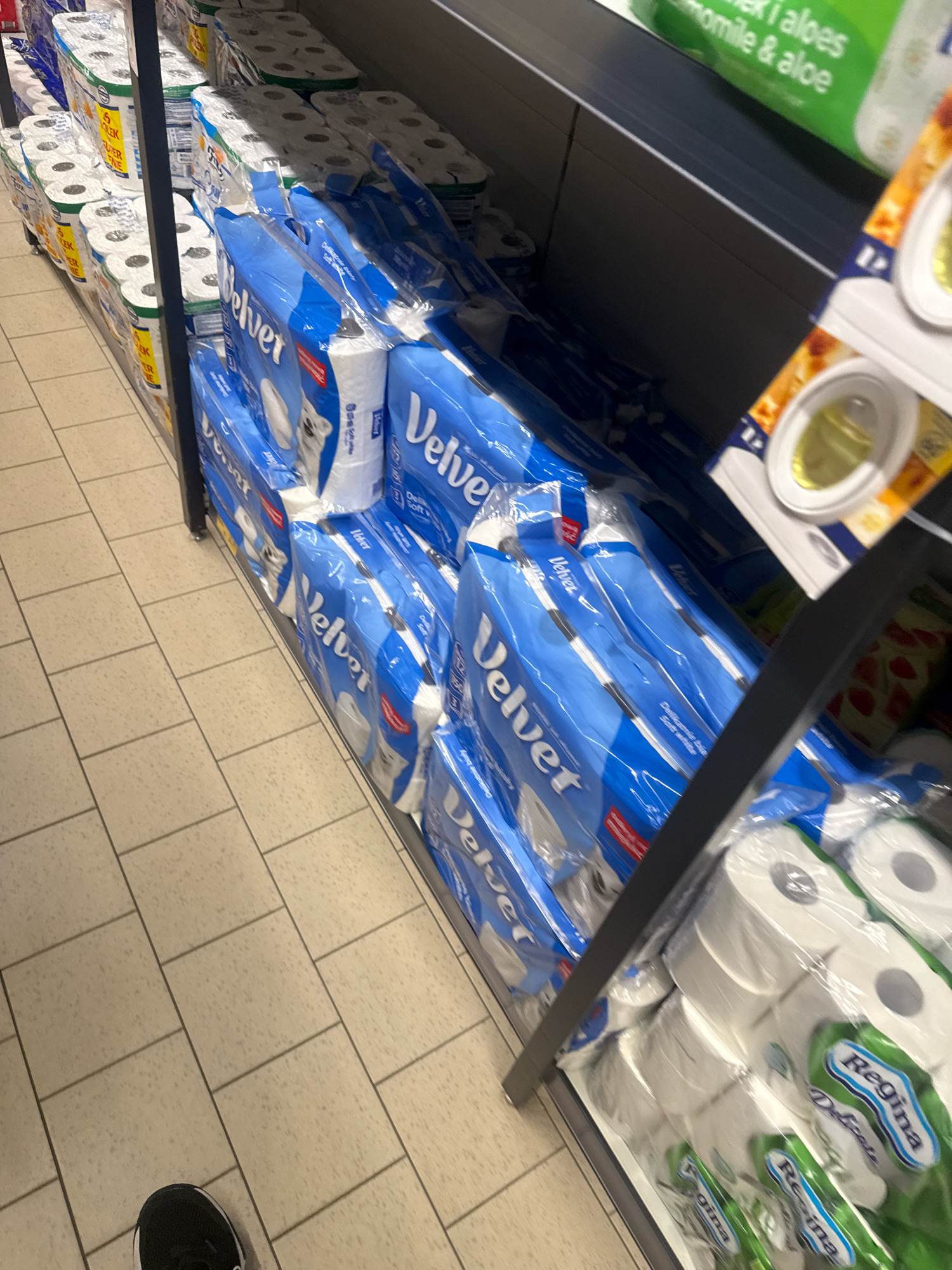 In Gorzow, Poland, on an aisle of a store, is a retail display of toilet paper. The toilet paper is packaged in blue and white plastic wrap with "Velvet" branding visible on some packs. There are multiple stacks of toilet paper. On a higher shelf, partially visible is a product with green packaging and text including "chamomile & aloe." To the right of the toilet paper stacks, on the same shelf, are packs of capsules with yellow contents. On the bottom shelf, more toilet paper is visible, with "Regina" branding on some packs. The floor is tiled with light-colored square tiles. A person's black and white sneaker is visible in the lower left corner of the image, indicating the perspective is from eye-level with someone standing in the aisle.