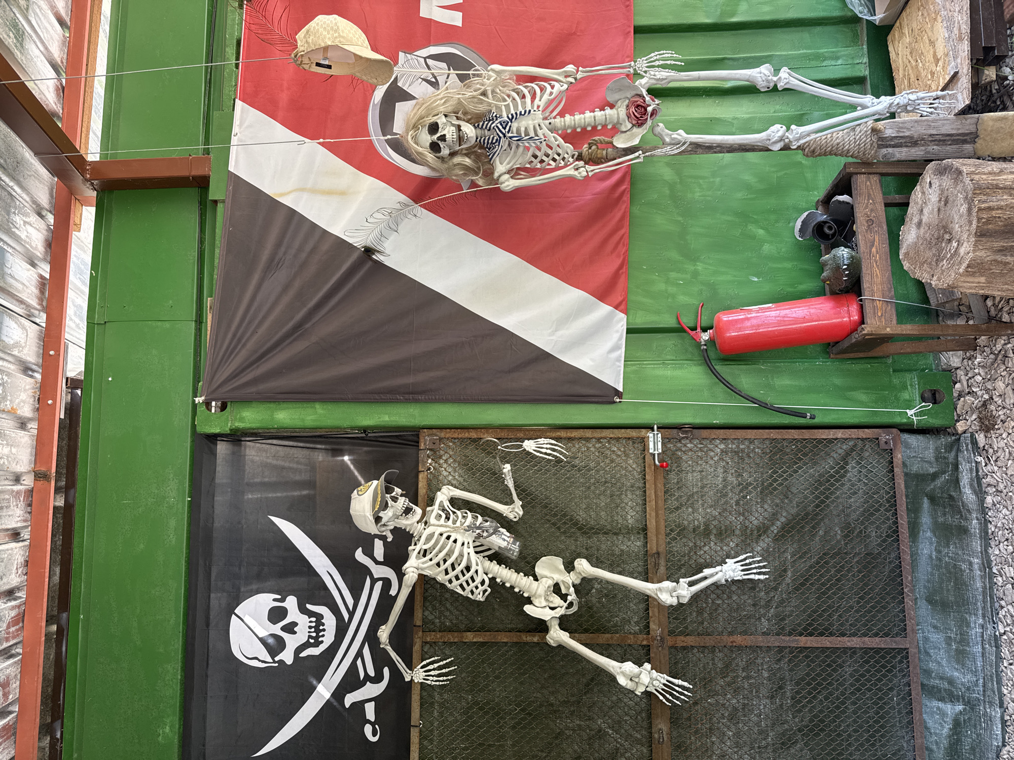 The image shows a vertical view of an outdoor Halloween display in Lukovit, Bulgaria. The scene is constructed against a green shipping container wall. On the upper section, a red, white, and black flag is displayed with a female skeleton adorned with a blonde wig, a striped bow tie, a straw hat, and a rose decoration. A rope appears to secure the skeleton. To the right of the flag is a red fire extinguisher and various wooden objects. Below, a black flag with a white skull and crossbones (pirate flag) is visible, and a male skeleton in a baseball cap is positioned in front of a wire mesh door, giving the impression that the skeleton is falling through the door. The scene is positioned on a gravel or stone surface.