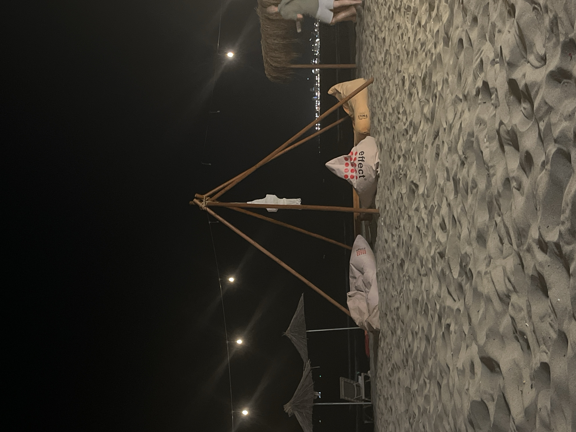 An outdoor nighttime scene on a sandy beach in Bashkia Durrës, Albania, is depicted. A temporary structure, possibly a canopy or shade, is made of wooden poles and adorned with string lights. In the foreground, several beanbag chairs are arranged on the sand. One beanbag has "effect" written on it. In the background, two straw umbrellas provide shade for beach chairs. The distant shoreline features lights from buildings and potentially a city. A person in a green shirt and white shorts is walking away from the camera towards the right side of the frame, near a thatched-roof structure. The sand shows footprints and undulations.