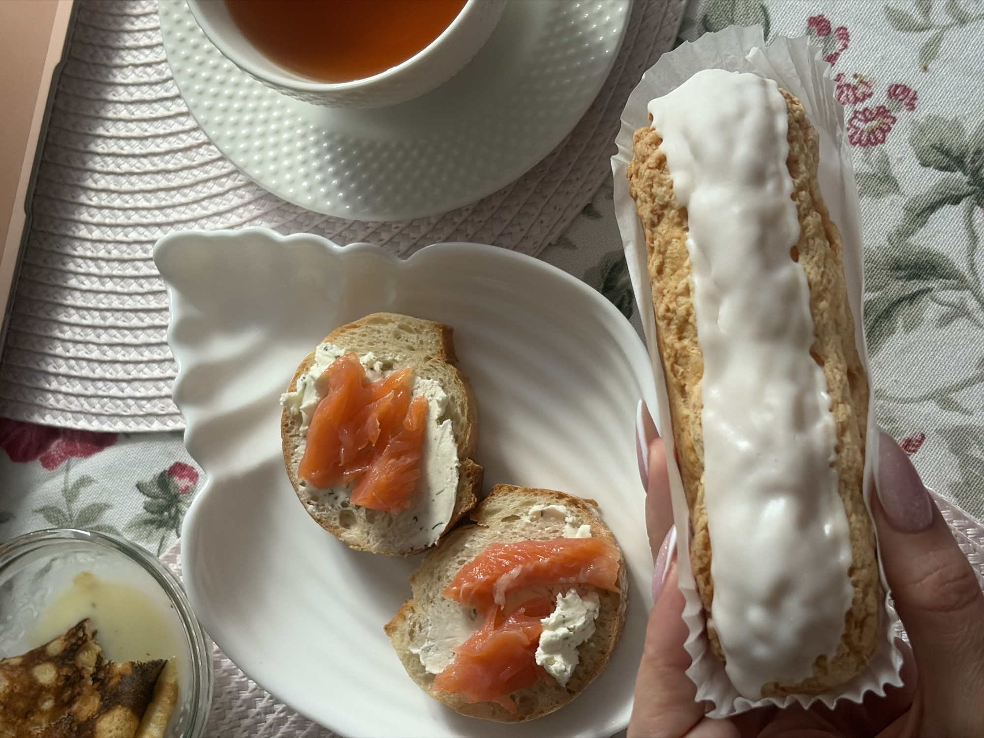 The image shows a table setting with several food items. A cup of tea sits on a saucer. A white plate holds two slices of bread topped with cream cheese and salmon. A clear glass bowl contains what appears to be a stack of crepes. In a person's hand is a pastry with a white icing strip on top, inside of a paper holder. The scene appears to be indoors. The tablecloth has a floral design. Without additional information, it's impossible to confirm that the location is Saint Petersburg, Russia.