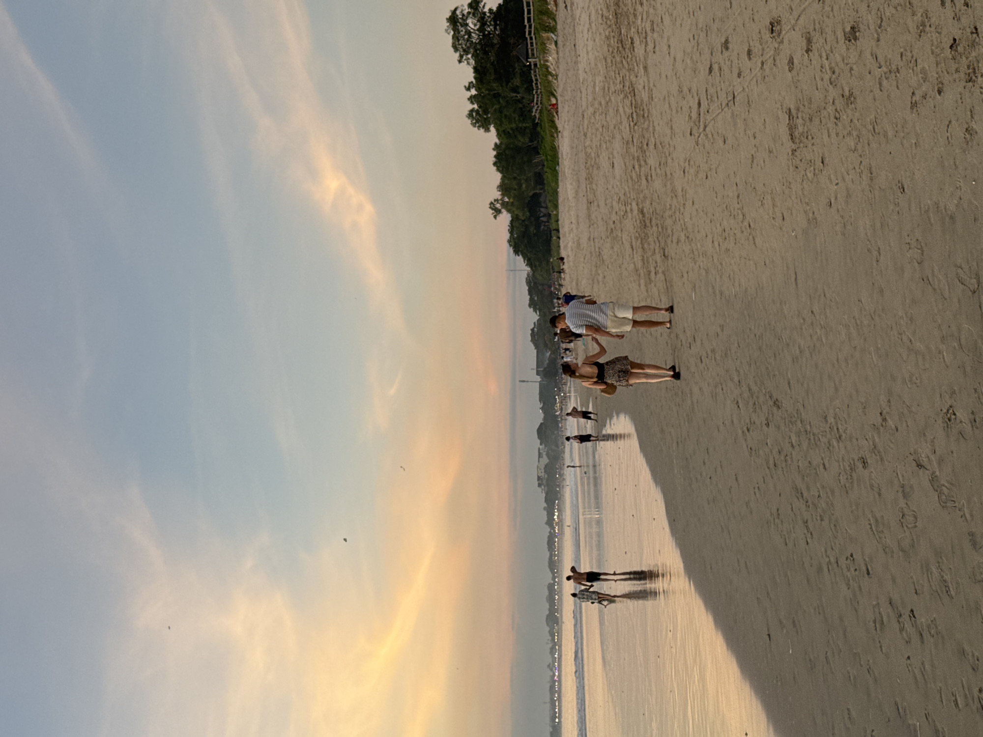 The image shows a beach scene at sunset, likely in Kuta Selatan, Indonesia given the prompt. Several people are walking along the shoreline. The sky is a mix of blue and orange hues. The beach itself appears sandy with footprints visible. Trees and possible structures are visible further inland, along the edge of the beach. Two figures are standing closer to the top of the image.