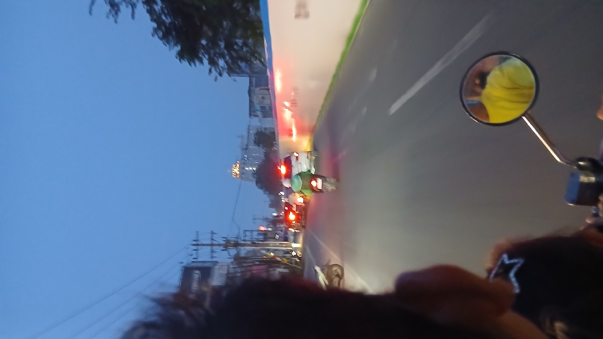 The image captures a street scene in the city of Medan, Indonesia, during dusk. The perspective is from a vehicle, likely a motorcycle, looking forward. In the foreground, the back of a person's head and shoulders are visible, wearing a dark shirt with a white star pattern. A motorcycle rearview mirror is also prominent, reflecting a person wearing a bright yellow t-shirt.
The street is lined with utility poles and buildings. Traffic is present, with several vehicles including cars and motorcycles. A motorcycle with a rider in a green jacket is in the middle ground, its brake lights illuminated. Another motorcycle is parked on the left side of the road. The road surface is asphalt, with white lane markings visible. A concrete wall with red lights and some graffiti runs along the right side of the street. In the distance, a tall building with illuminated signage can be seen. The sky is a deep blue, indicating twilight. The overall scene suggests a busy urban environment at the end of the day.