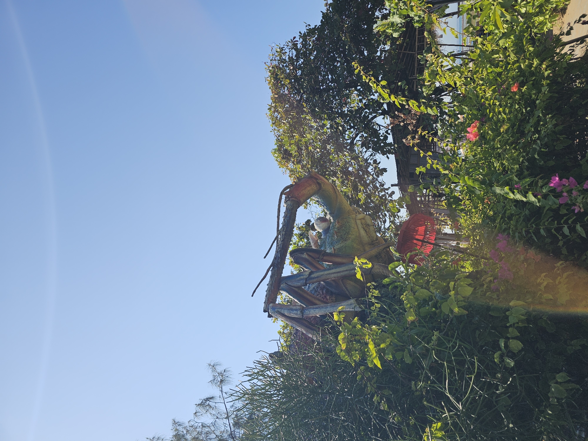 This is an outdoor, low-angle shot of a large, colorful sculpture of a lobster or crustacean, partially obscured by lush green foliage. The sculpture is predominantly green and blue with red accents on its claws and antennae. It appears to be part of a park or tourist attraction. In the background, there is a thatched-roof structure, possibly a hut or a small building, and beyond that, a glimpse of the ocean under a clear blue sky. The sun is bright, casting lens flares across the image. The foliage includes various types of plants, some with small pink and purple flowers. The overall impression is of a tropical setting, possibly in Denpasar, Indonesia, known for its beaches and cultural attractions. There are no people visible in the image.