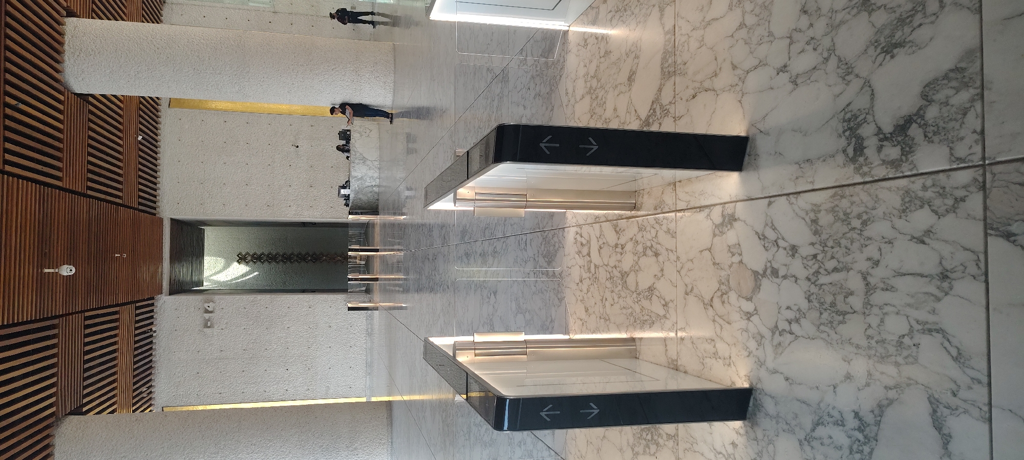 The image shows an interior view of a building, potentially a museum or gallery, located in Mexico City, Mexico. The floor is covered in large marble tiles. Directional signs with black backgrounds and white arrows are prominently placed. People are visible in the distance, their presence suggesting the building is open to the public. One person is looking at their phone. The ceiling features a modern wooden slat design. A large window or opening offers a glimpse of a textured wall and geometric design within. The overall lighting is bright and appears to be a combination of natural and artificial sources.