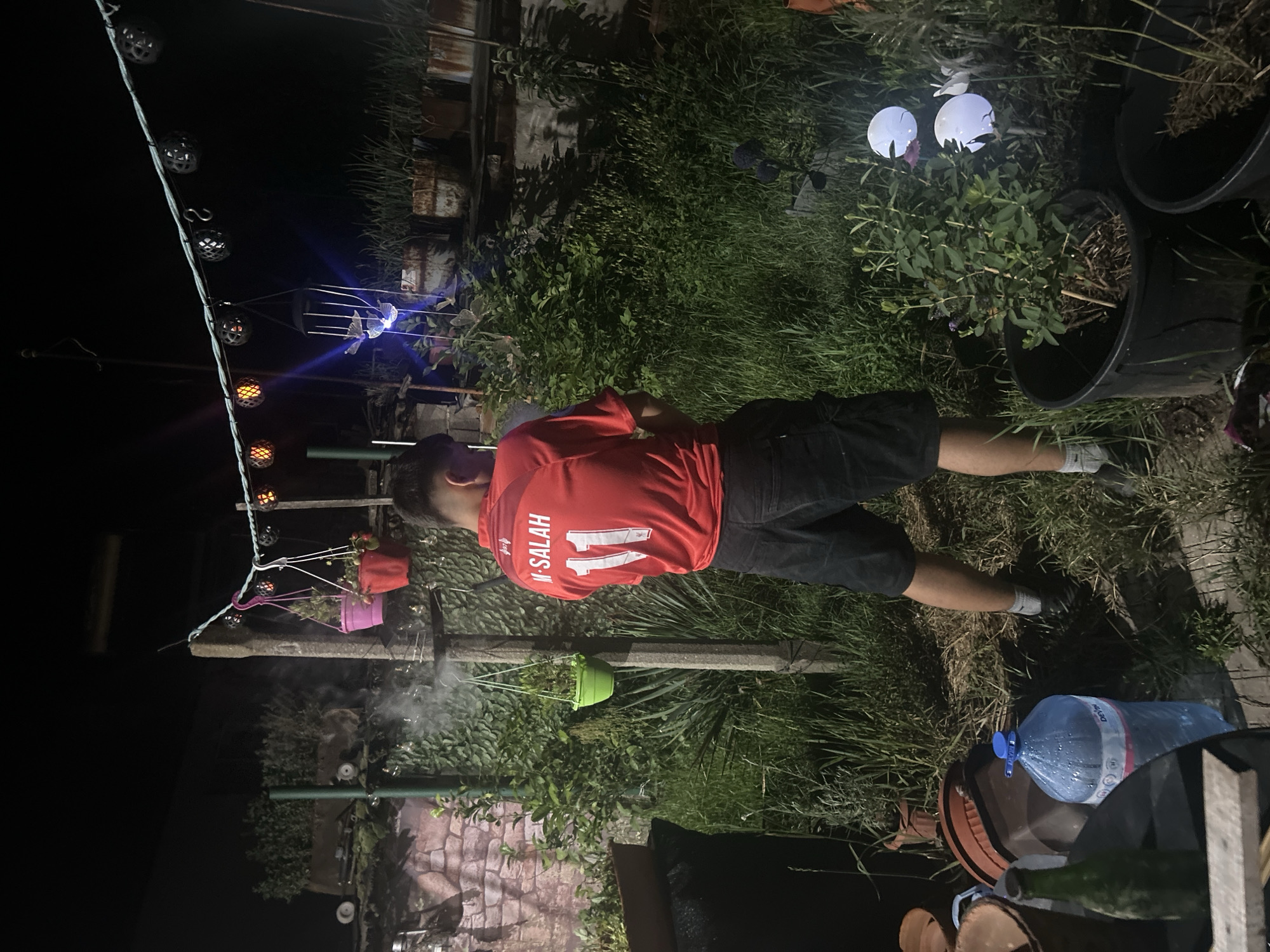 A man wearing a red football jersey with "M. SALAH" and the number "11" on the back, along with black shorts and white socks, stands facing away from the camera in a dimly lit outdoor setting that appears to be a backyard or garden. He is positioned among tall green grass and plants. In the foreground, to the left, is a table with a green glass bottle and a large blue water bottle. Hanging above, strung between posts, are a series of decorative lights, some emitting a warm glow, and several hanging planters with plants. To the right, in the background, are more potted plants and some rusty cylindrical containers, possibly planters as well. The overall scene suggests a casual, possibly evening gathering in a residential outdoor space.