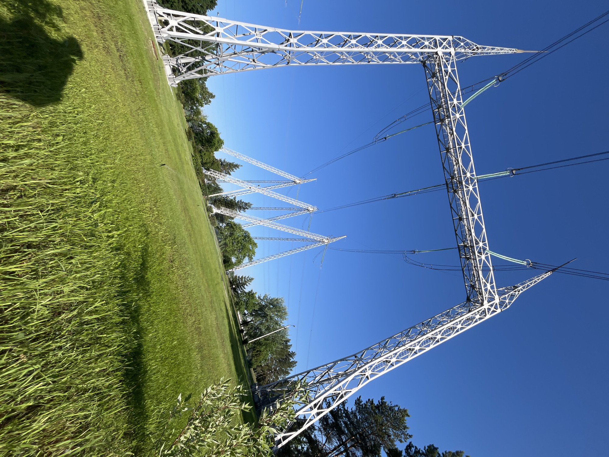 The image shows a bright, sunny day in Chaikovsky, Russia, with a clear blue sky. In the foreground, a lush green grassy hill slopes downwards. On the left side of the hill, a large, white metal electricity pylon is prominently featured, with its lattice structure supporting overhead power lines. Further in the background, a series of similar, though smaller, electricity pylons are visible marching across the landscape. These pylons are also white and have a distinctive angular design, with their V-shaped structures contrasting with the broader A-frame of the pylon in the foreground.
The pylons are spaced out across the hill and appear to be part of a transmission line. Trees, a mix of deciduous and coniferous, dot the landscape behind the pylons, suggesting a semi-rural or park-like setting. The grass on the hill is vibrant green and appears to be well-maintained, with some shadows from unseen objects, possibly including the photographer, visible on the lower part of the hill. The overall scene depicts a clear, crisp day with industrial infrastructure integrated into a natural environment.